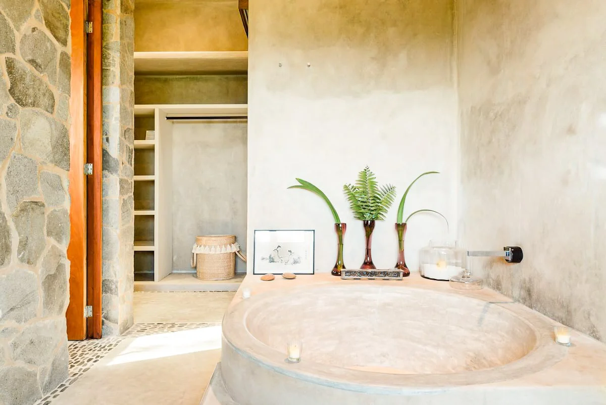 A rustic bathroom with a large oval stone bathtub decorated with green plants, candles, and a framed picture, featuring stone and plaster walls and a walk-in closet in the background.