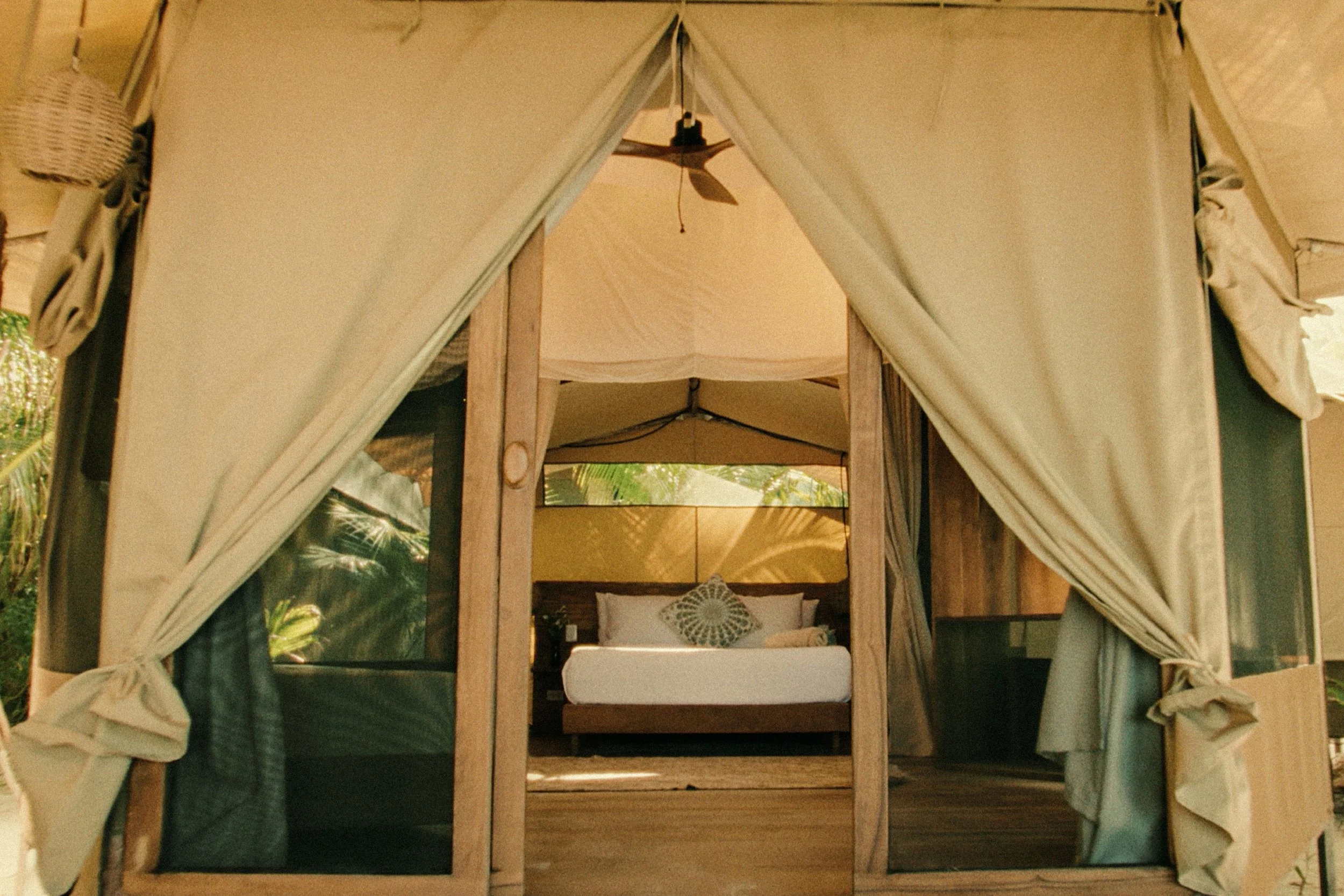 View of a luxurious bedroom inside a tent with large opening, wood accents, and tropical surroundings.