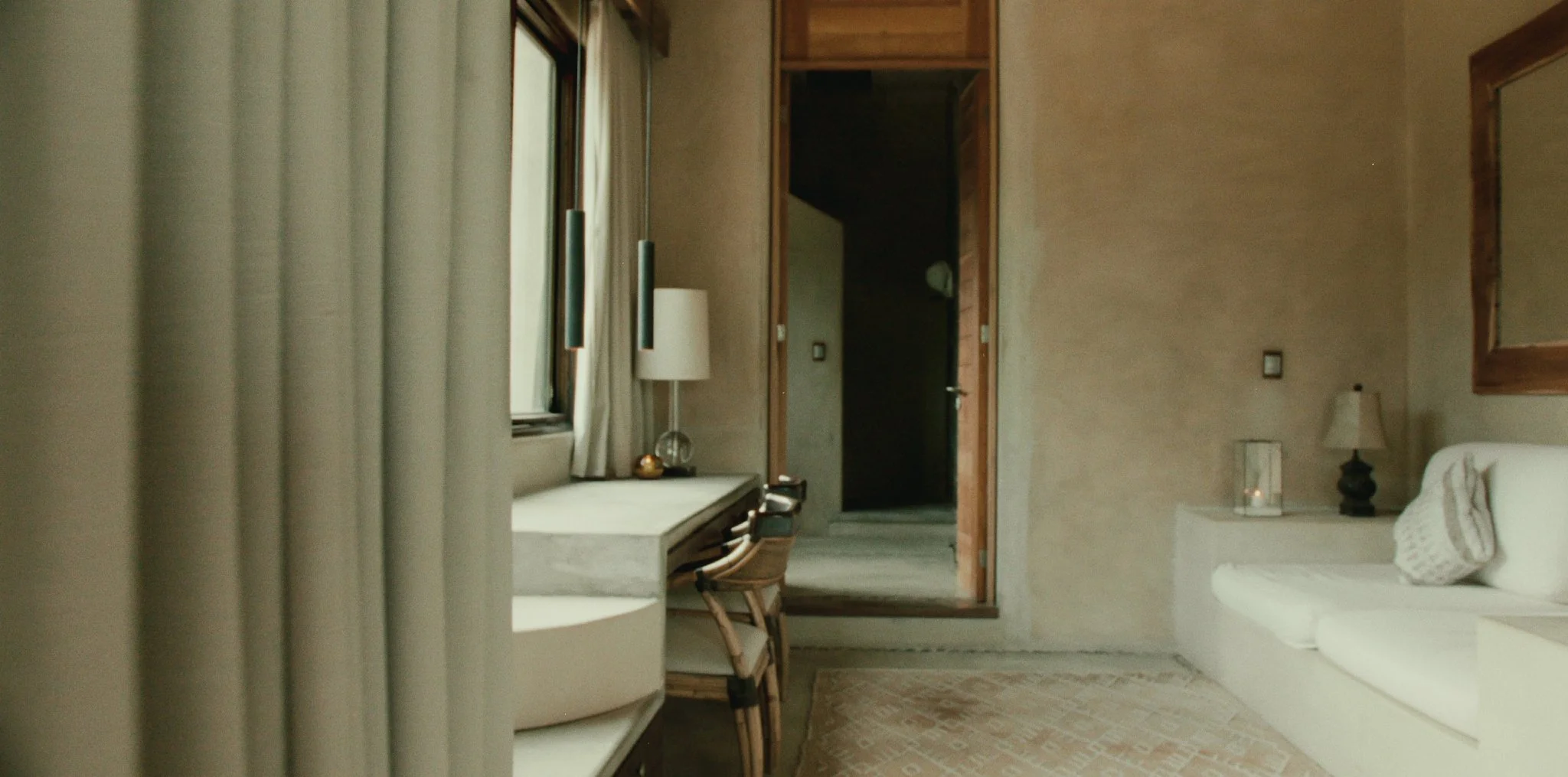 A cozy hotel room with beige walls, a white couch with a patterned pillow, a small desk with a wooden chair, a window with beige curtains, a table lamp, a framed mirror, and a small nightstand with a candle.