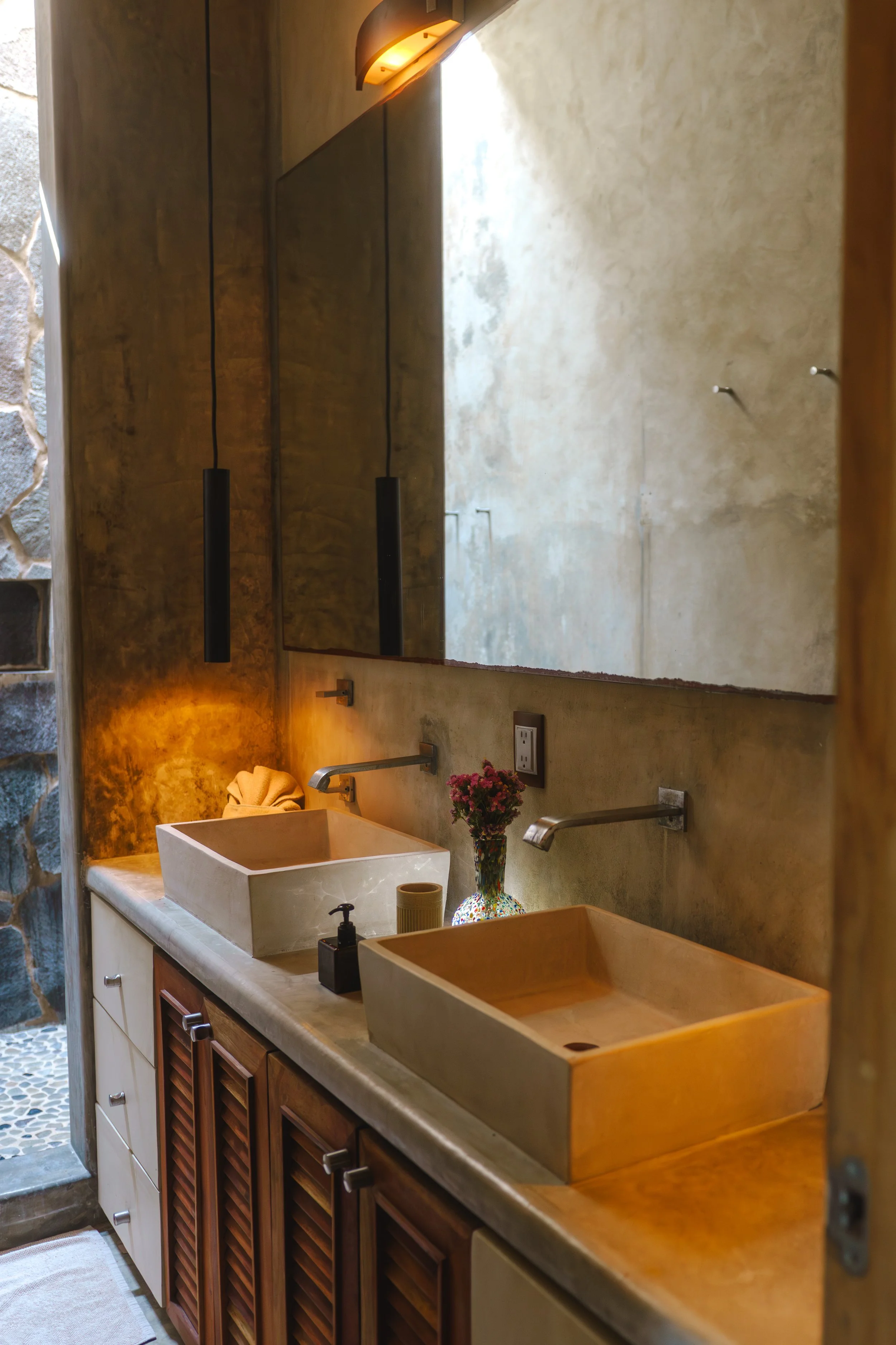 A bathroom vanity with two rectangular vessel sinks, a large mirror, and decorative accessories including a vase with flowers, a soap dispenser, and a towel. The wall behind the sinks is textured with a warm tone, and there are two modern pendant lig