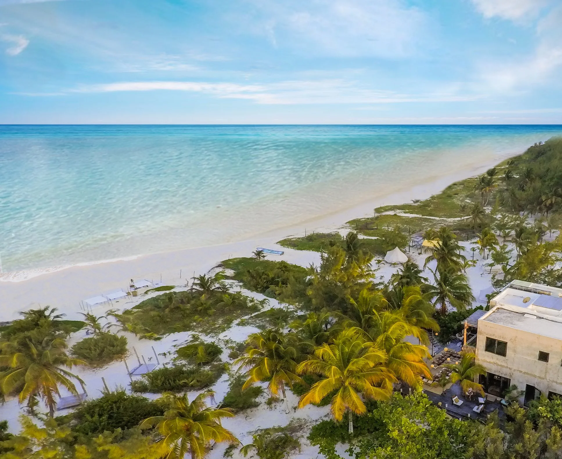 Aerial view of a tropical beach with white sand, turquoise water, lush green palm trees, and a few structures along the shoreline.
