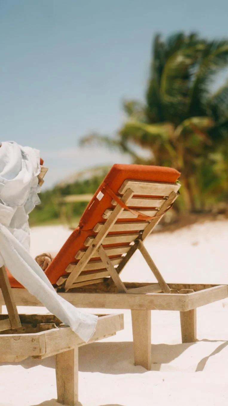 Wooden beach lounge chair with orange cushion on white sandy beach, with white cloth draped over nearby chair and palm trees in the background under a clear blue sky.