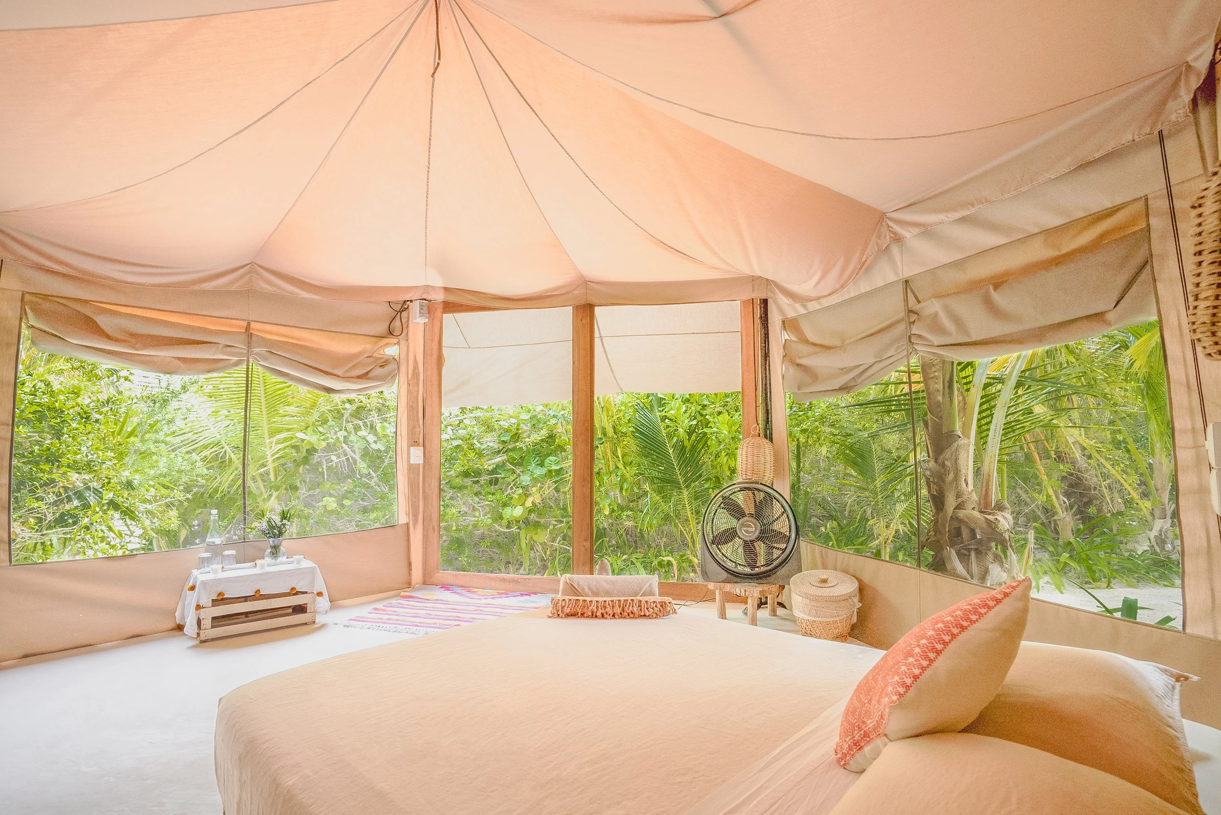 Room with a large bed, surrounded by windows with green tropical foliage outside, beige canopy ceiling, and simple decor.