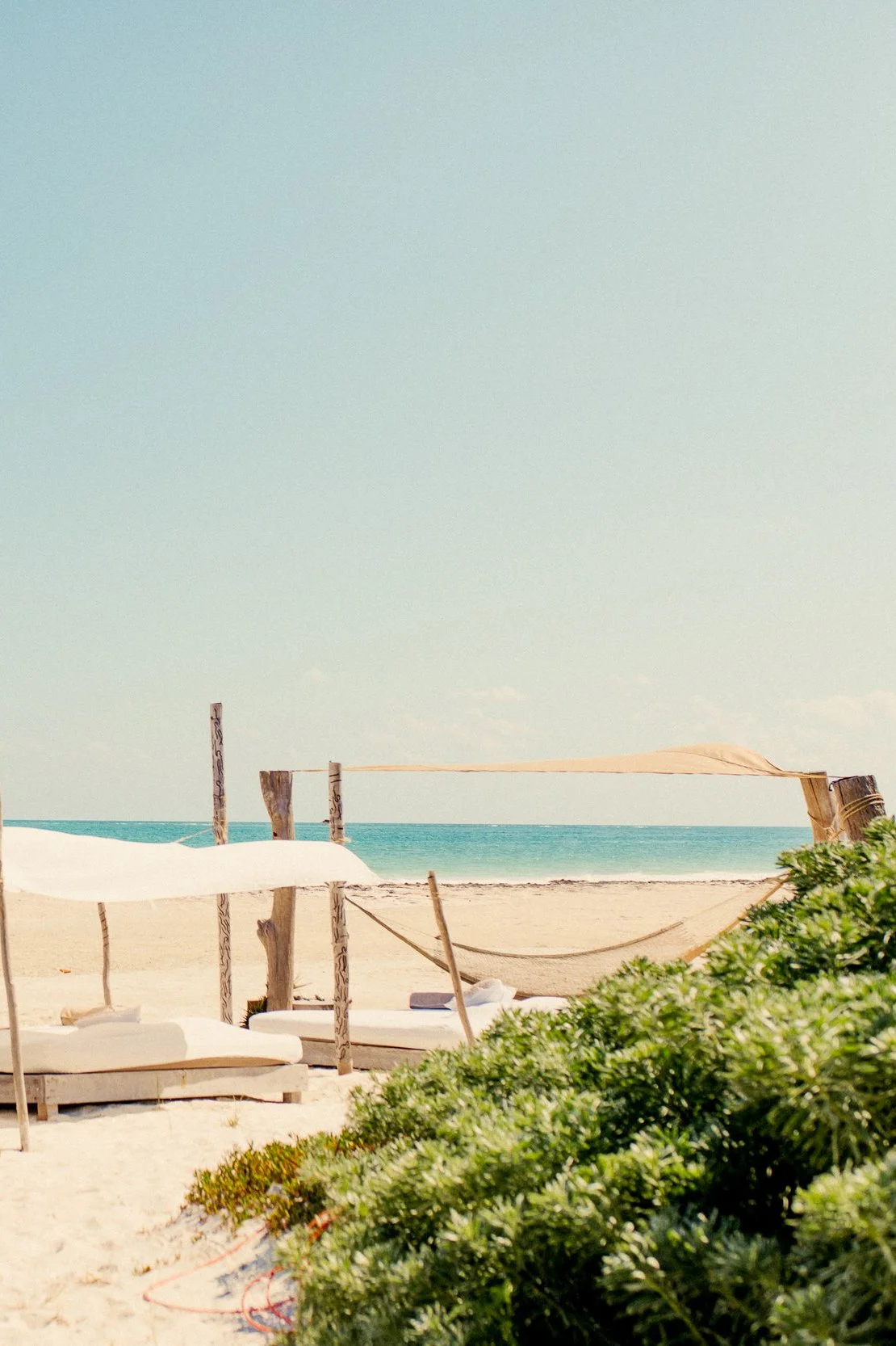 Empty beach with lounge chairs and a canopy, ocean in the background, clear sky, and some green bushes in the foreground.