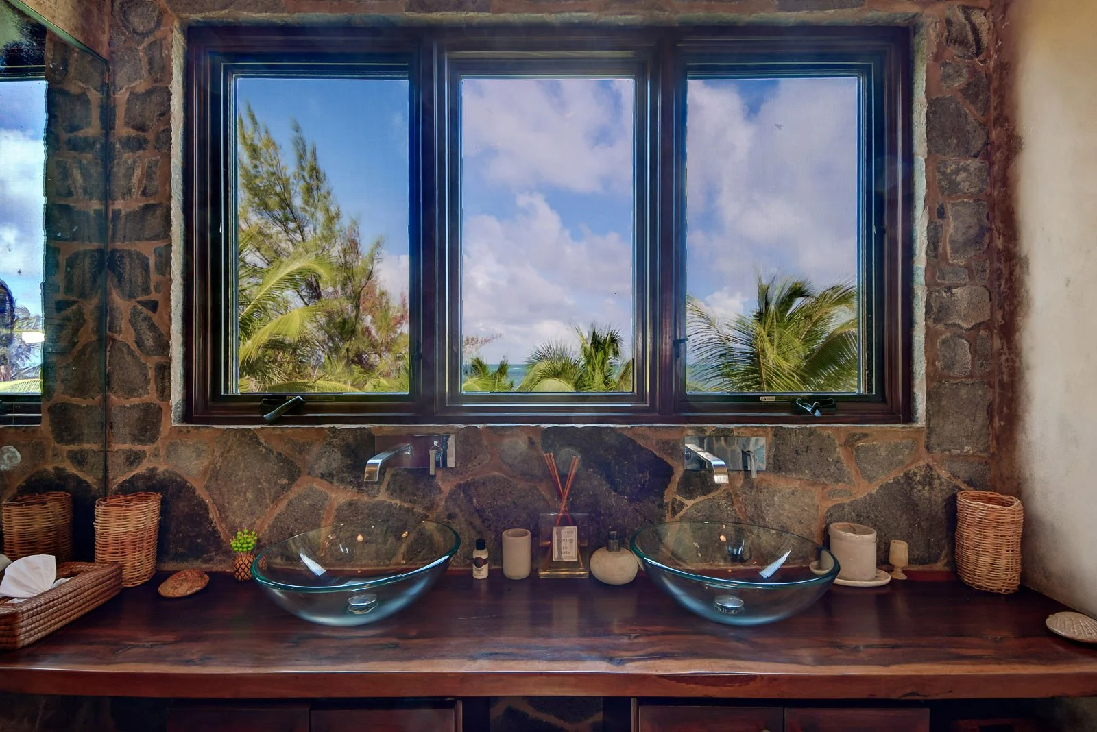 Bathroom with two glass sinks on a wooden counter, a window showing a tropical view with palm trees and blue sky, and various personal care items on the counter.