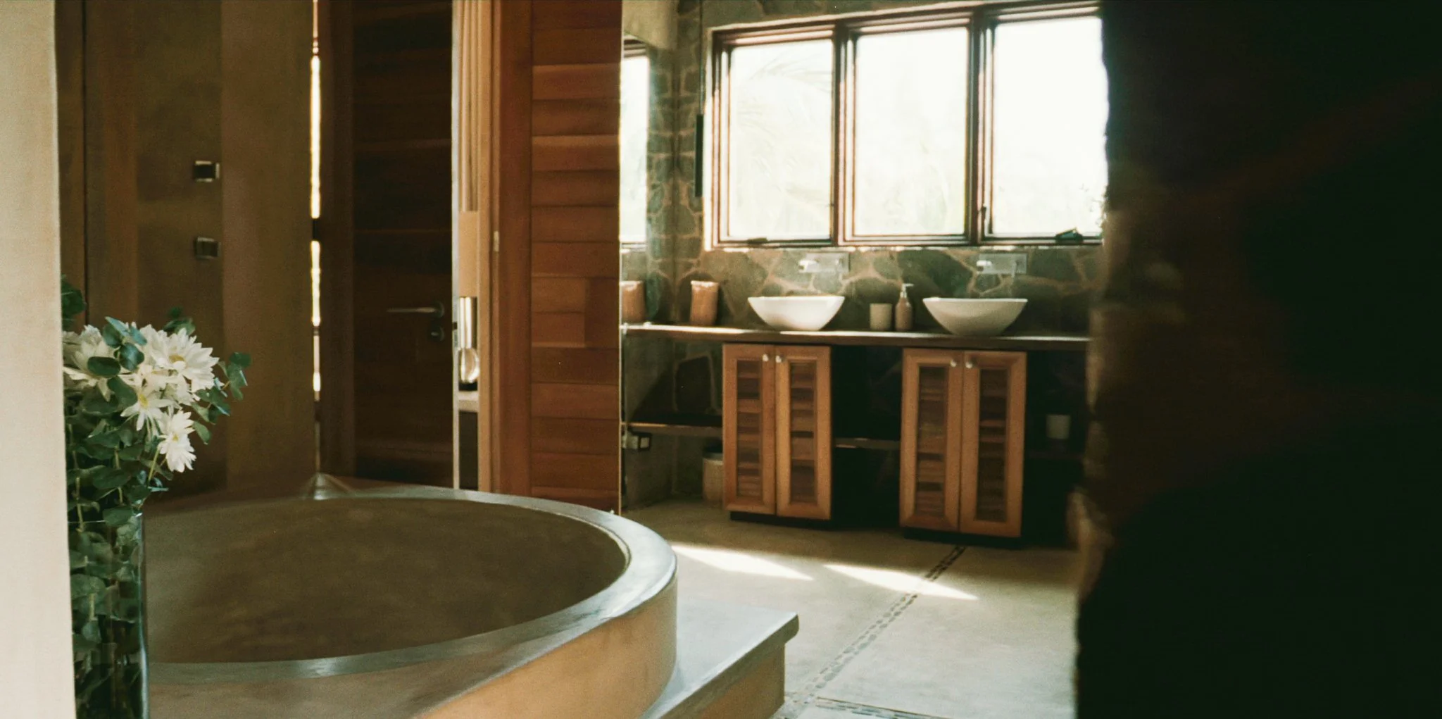 A bathroom with wooden cabinets, two white vessel sinks, large windows, and a stone wall in the background, illuminated by natural light.