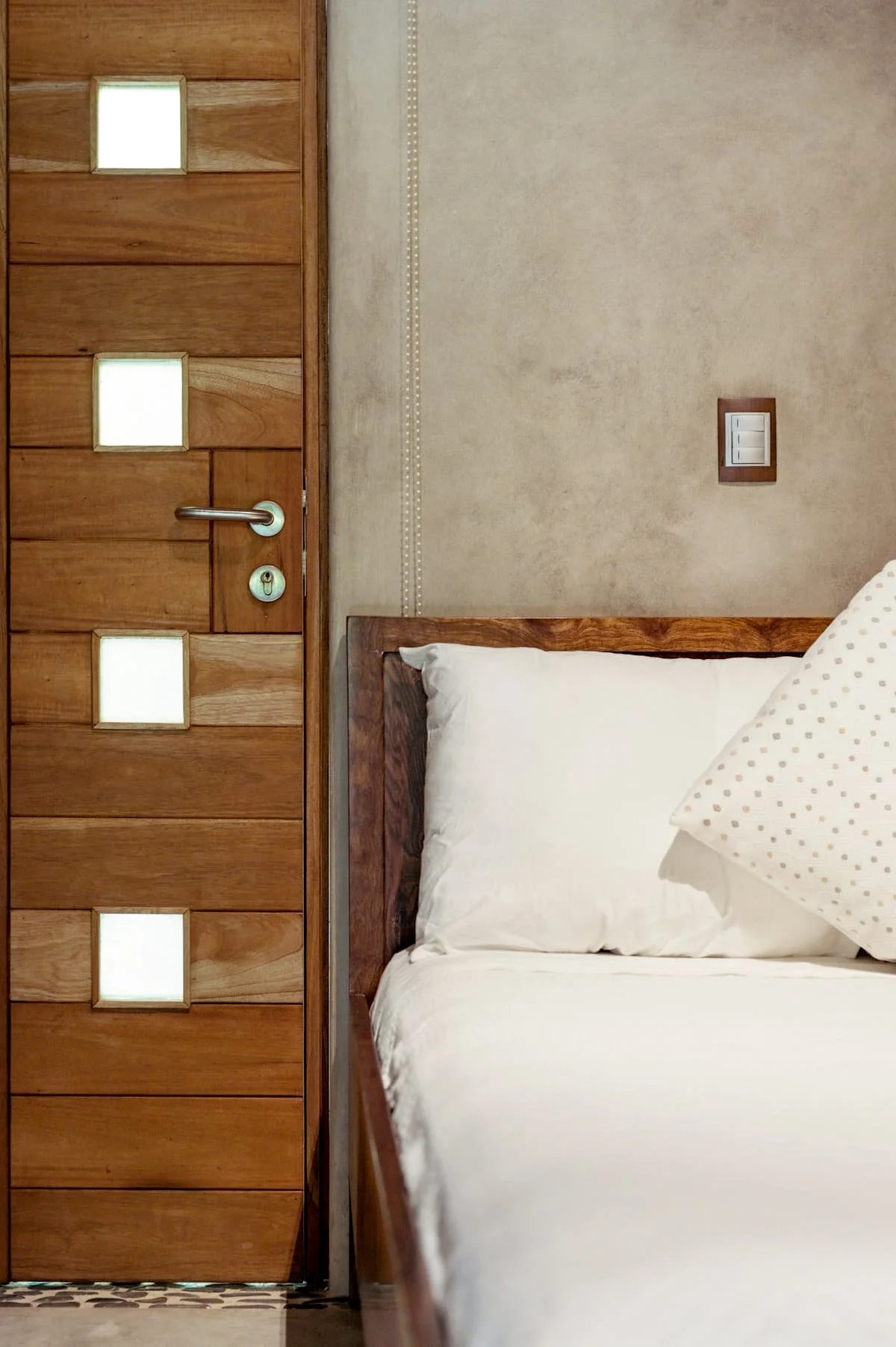 Close-up of a wooden door with square cutouts and a silver handle, adjacent to a bed with white linens and a polka-dot pillow, in a room with beige walls and a switch on the wall.