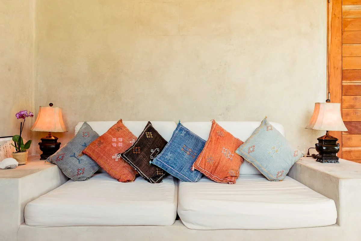 White sofa with seven colorful embroidered pillows, two table lamps, an orchid in a pot, and a wooden wall on the right side.
