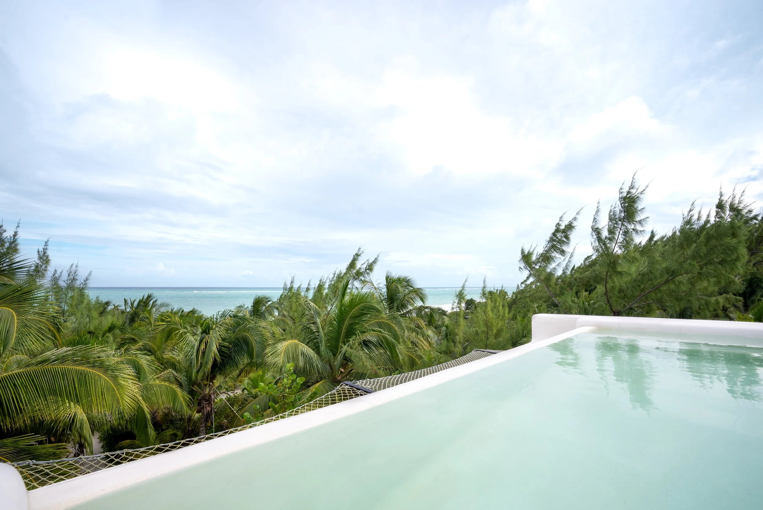Hot tub overlooking a tropical beach with lush green trees and the ocean in the background under a cloudy sky.