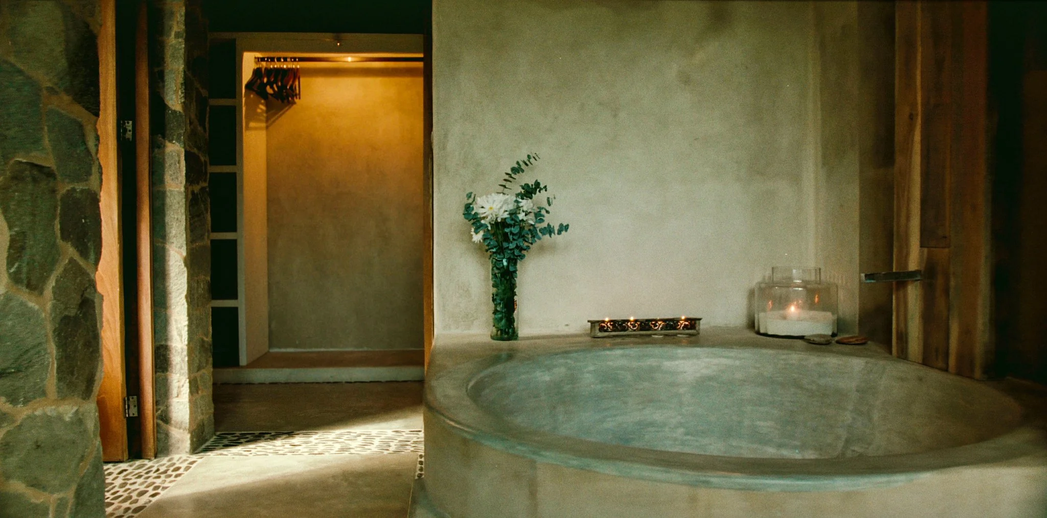 Bathroom with a stone and wood wall, a bathtub, a vase with white flowers, candles, and a towel warmer in the background.