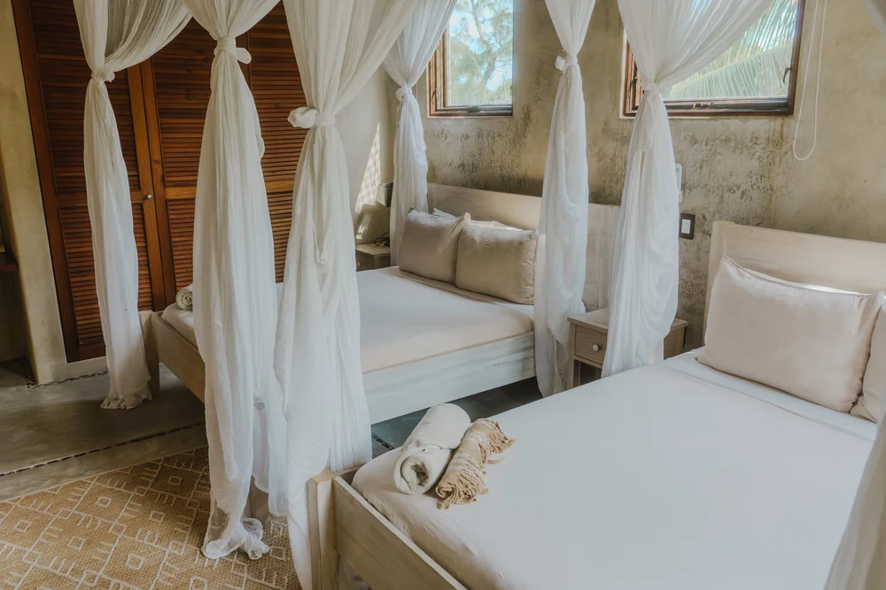 Two beds with beige linens and pillows in a rustic bedroom with at a luxury yoga and wellness retreat a shower curtain canopy in front of each bed. Towels are folded on one bed, and a window with wooden shutters is visible on the wall.