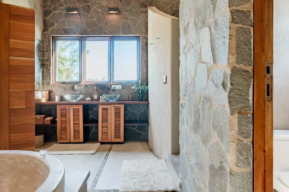 A rustic bathroom featuring a stone wall and window above a wooden vanity with two glass vessel sinks, baskets on the floor, and a view of palm trees through the window.