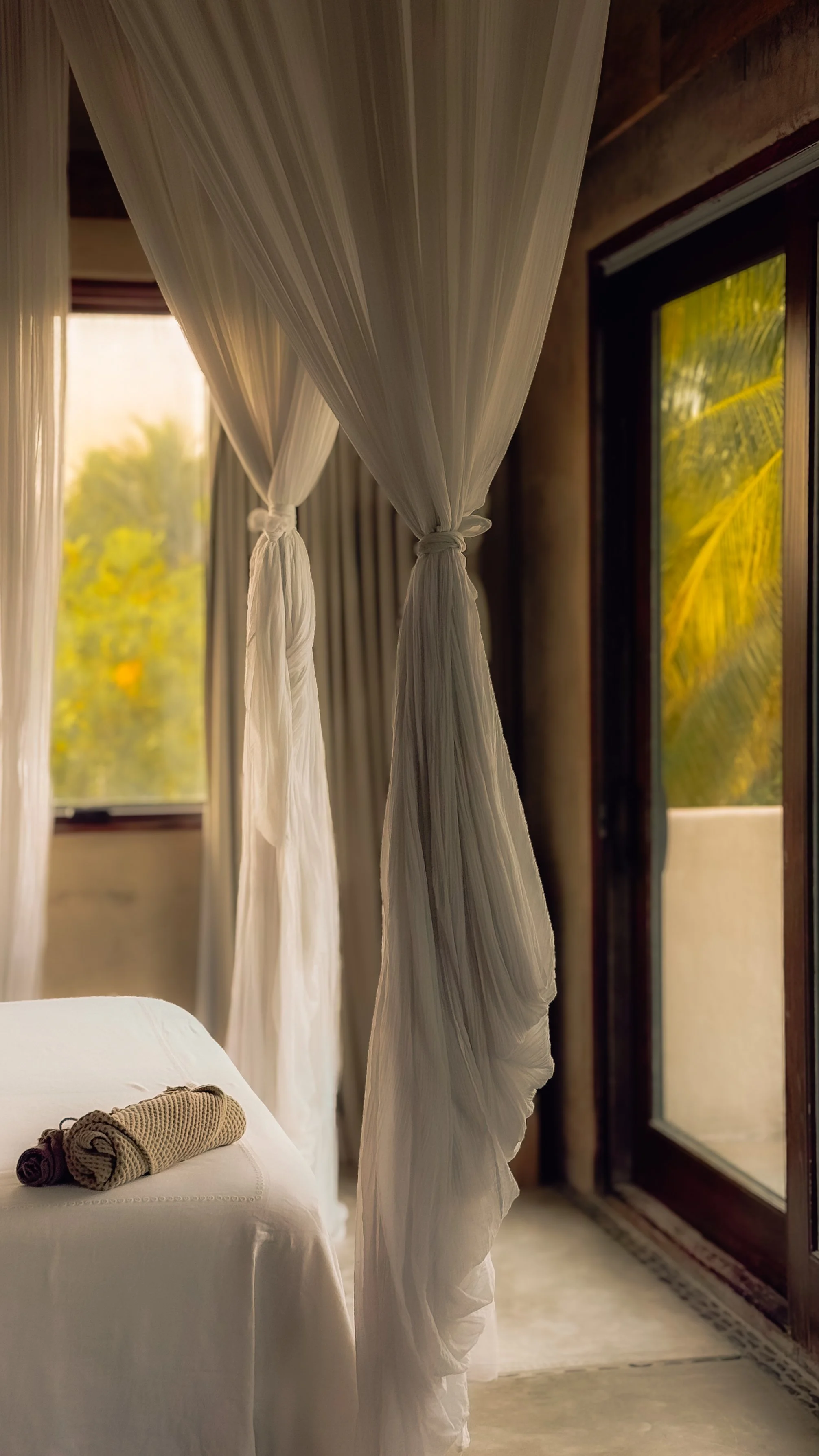 A bedroom with sheer white curtains tied back near a window, with a view of greenery outside. A bed with white linens has neatly rolled towels on it, and a sliding glass door opens to a balcony or outdoor space.