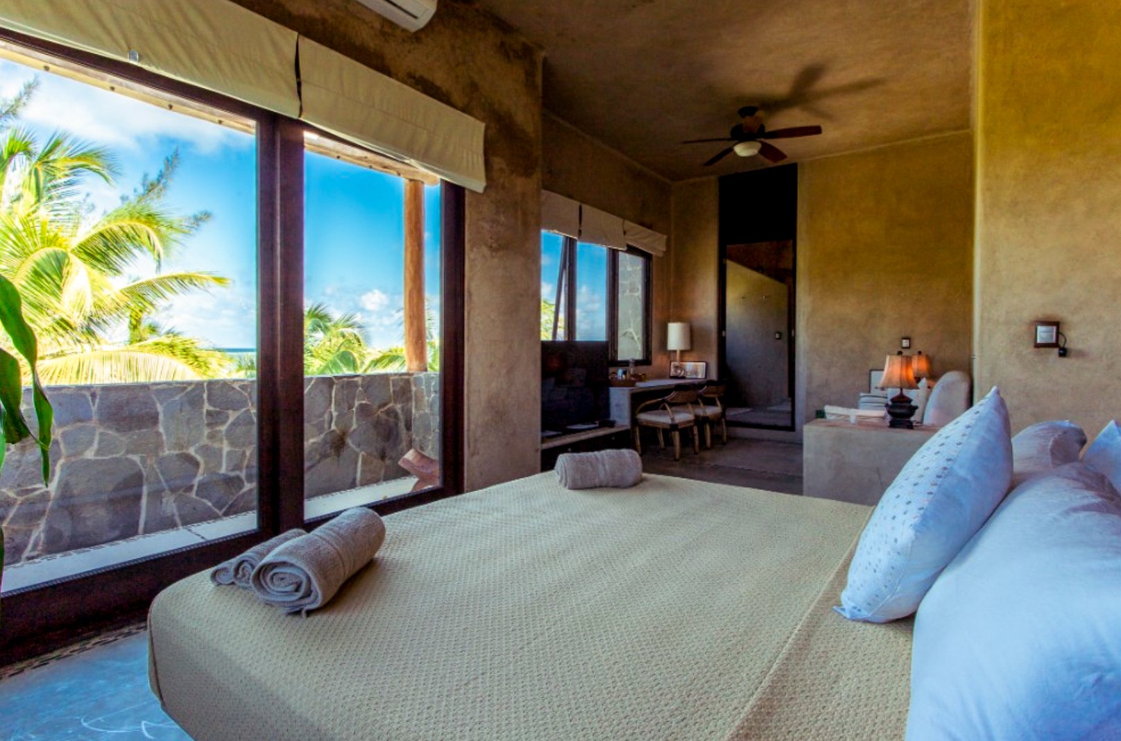 A bedroom with a large bed, white pillows, and rolled towels, overlooking a balcony with tropical palm trees and a blue sky outside.