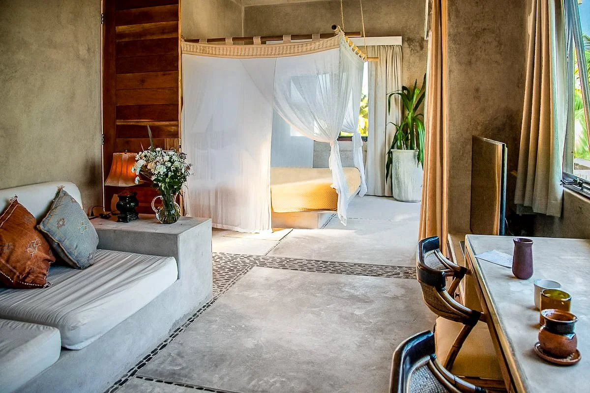 A cozy hotel room with a seating area, a bedside table with flowers and a lamp, a four-poster bed with a mosquito net, a potted plant, a television, and a dining table with cups and vases. Bright natural light filters through open windows with curtai