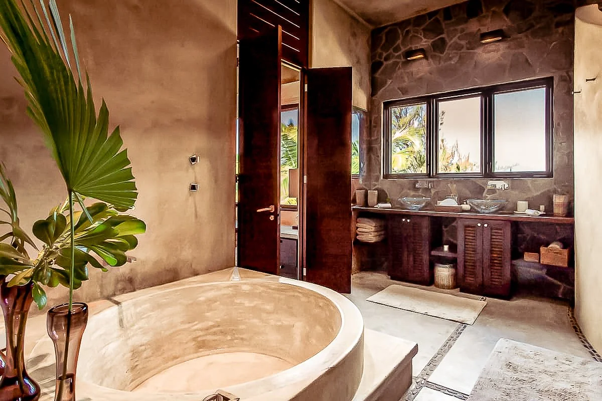 Bathroom with a large round bathtub, a plant in a vase, a wooden door, a window showing outdoor greenery, and a dark vanity with two sinks and shelves underneath.