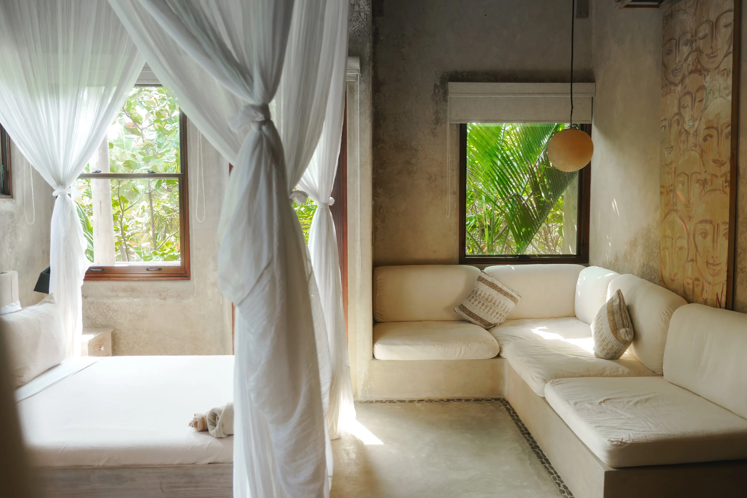 Sunlit bedroom with white curtains tied back, beige walls, a cream sectional sofa with patterned pillows, and windows showing green foliage outside.