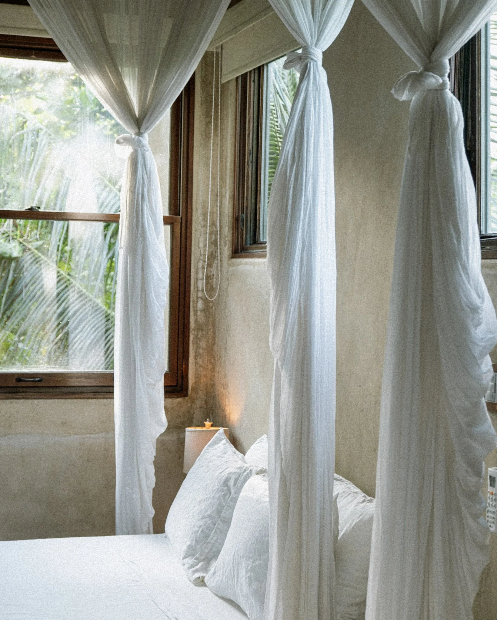 A cozy bedroom with white bed linens and a mosquito net canopy hanging from the ceiling, near open windows with greenery outside.