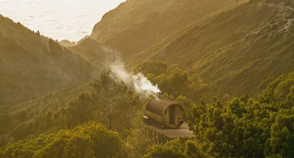 Wood-fired barrel sauna sending smoke across Amaria’s coastal valley at dusk, near Odeceixe