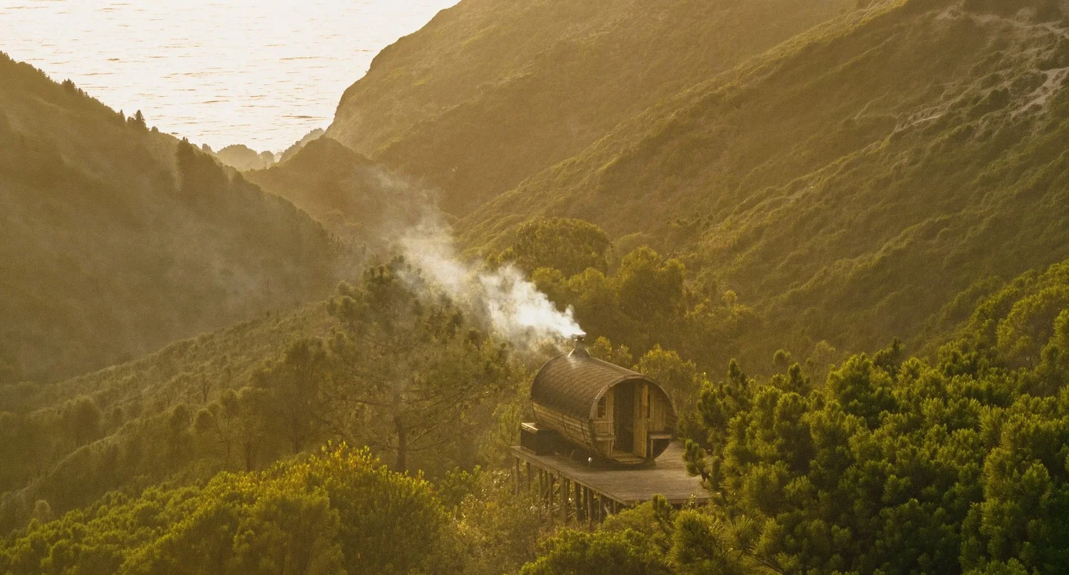 Wood-fired barrel sauna sending smoke across Amaria&rsquo;s coastal valley at dusk, near Odeceixe, Portugal
