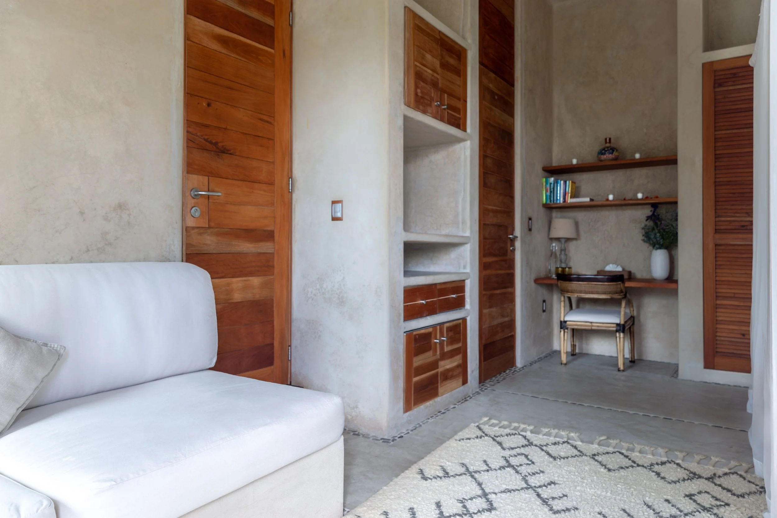 Interior view of a room with a white sofa on the left, wooden doors, built-in shelves, a desk with a chair, a lamp, books, and decorative items on the right, and a patterned rug on the floor.