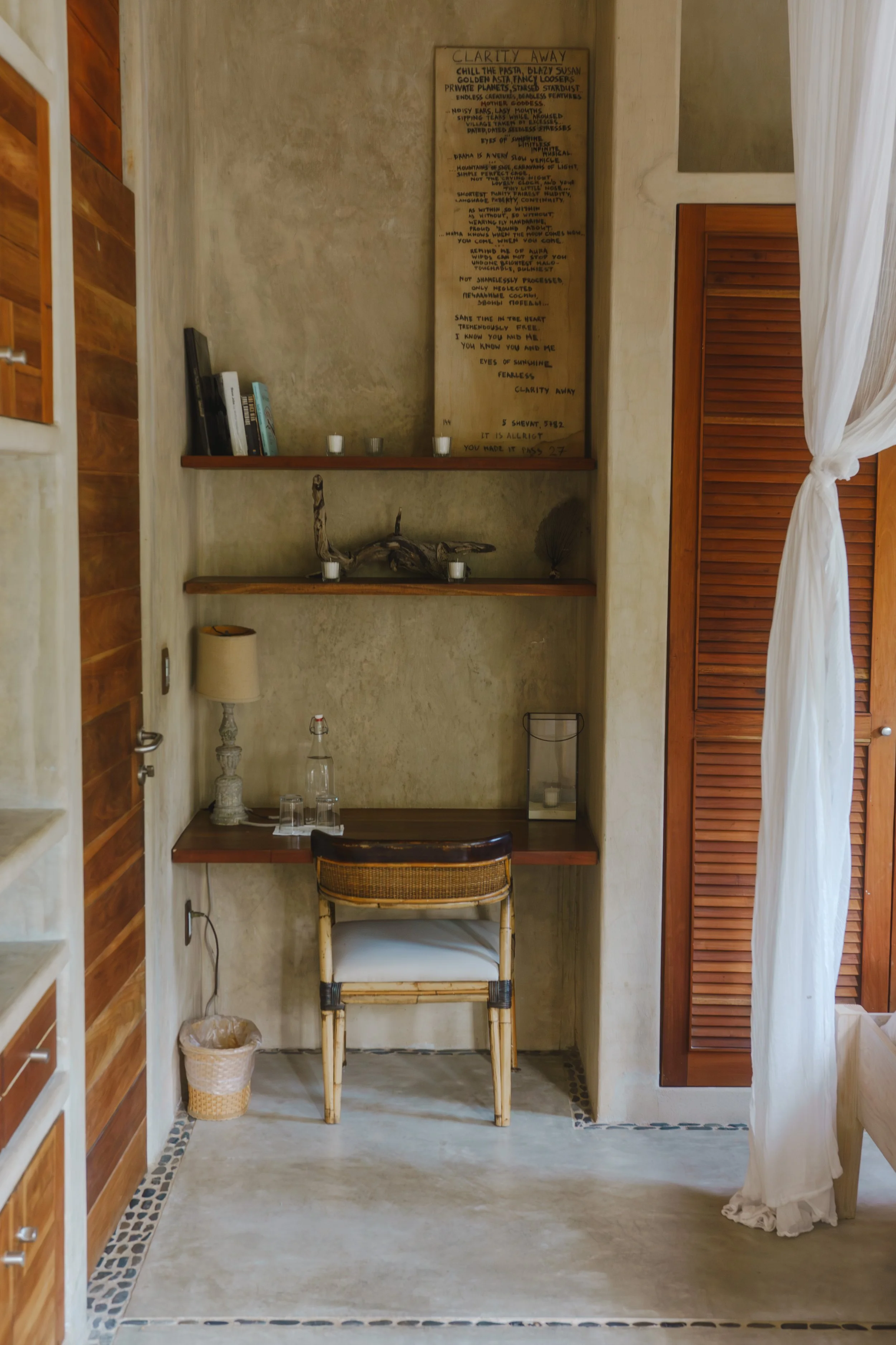 Small workspace with a wooden desk, rattan chair, beige walls, wooden shelves, a table lamp, and a curtain partially covering a wooden closet door.