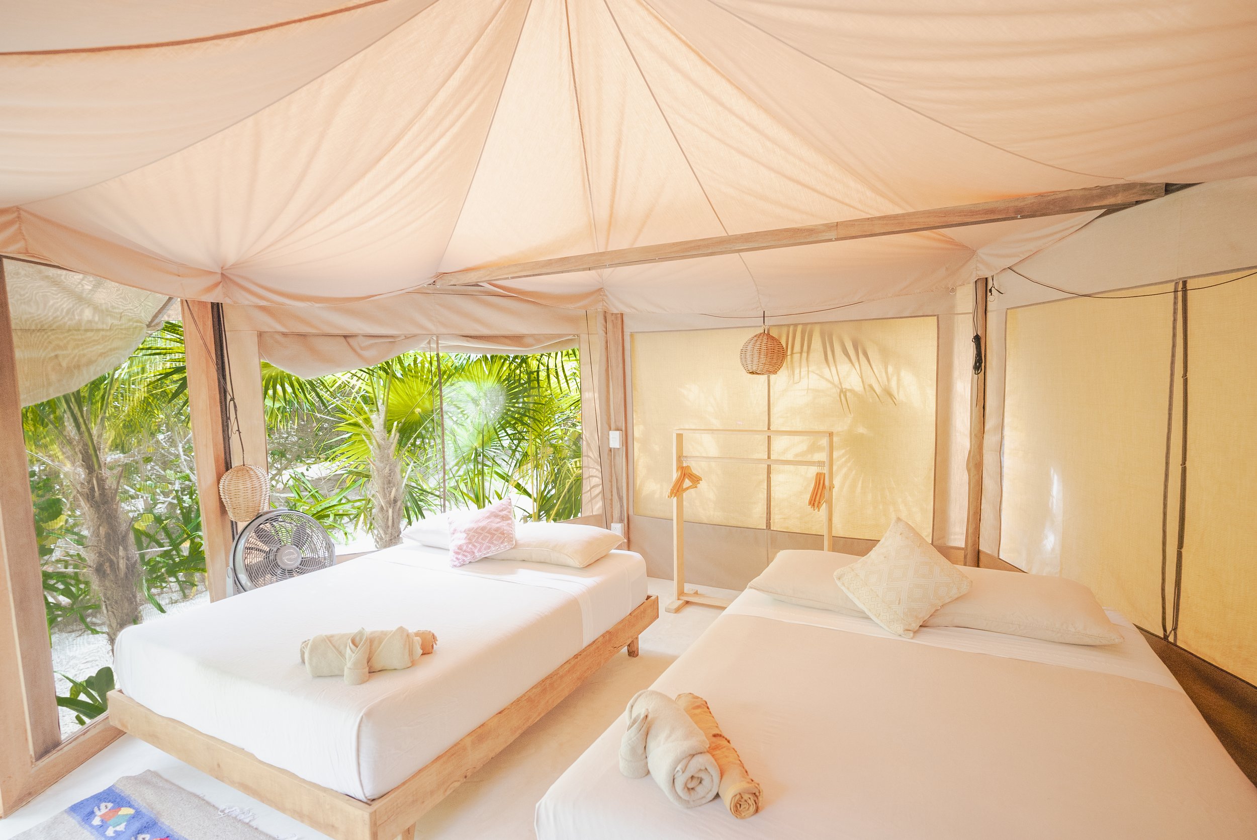 Two single beds with white linens and towels shaped like swans, inside a canvas tent with lush tropical plants visible outside.
