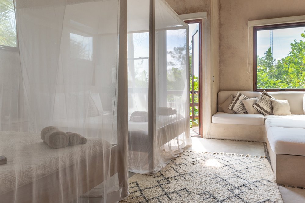 Bedroom with a groomed bed behind a white mosquito net, beige walls, a rug, and a window showing green trees outside.