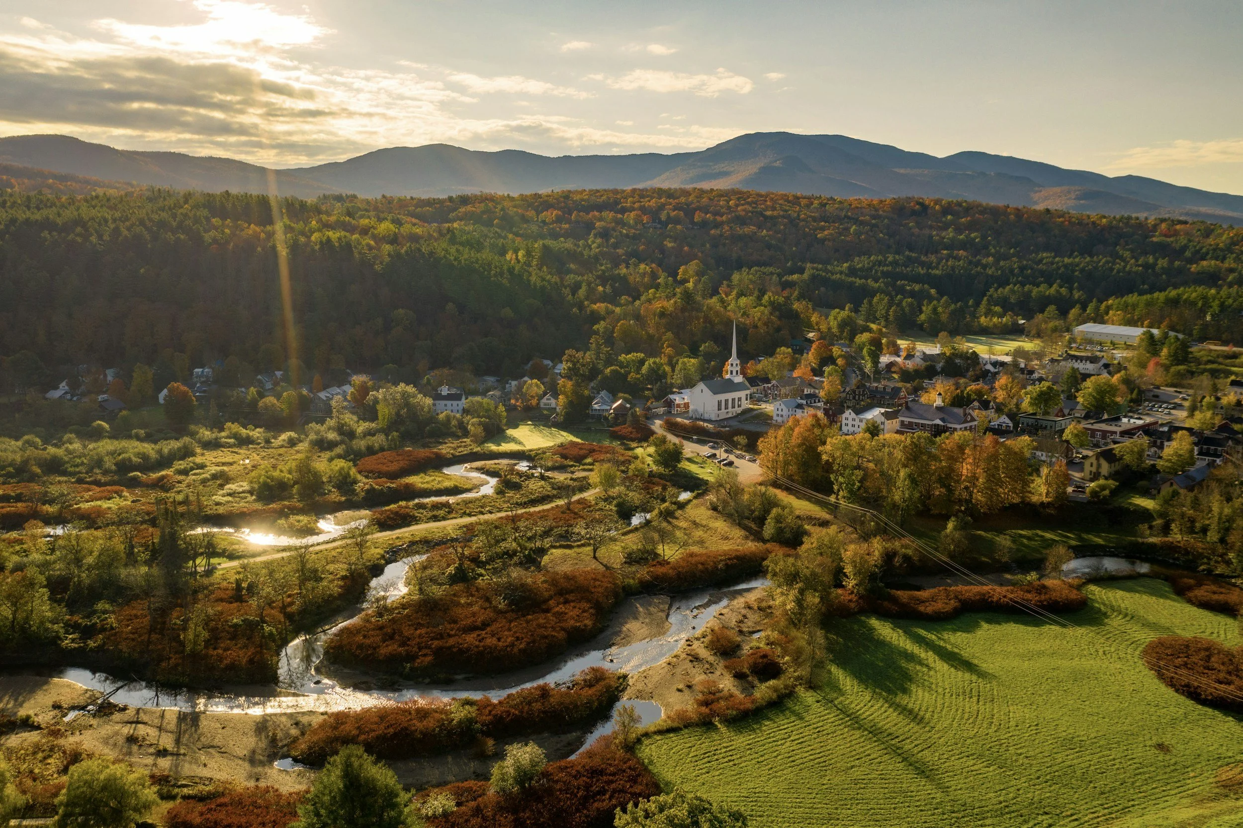 Aerial view of a small town surrounded by lush autumn foliage, with a church and winding river in the foreground, and mountains in the background at sunset.