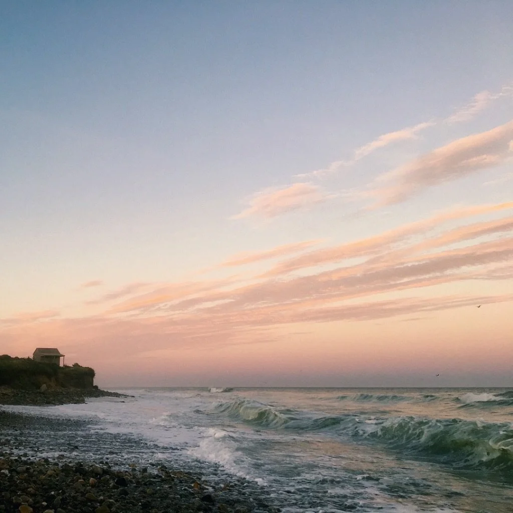 A beach at sunset with waves crashing onto a pebbly shore, a small house on a cliff in the distance, and a pastel-colored sky with clouds.