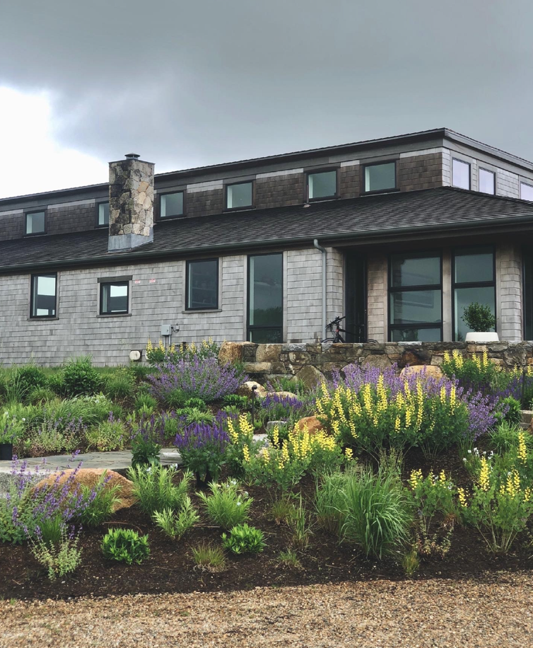 A modern two-story house with a garden in the front yard filled with colorful purple, yellow, and green plants. The house has gray shingles, a large stone chimney, and multiple windows.