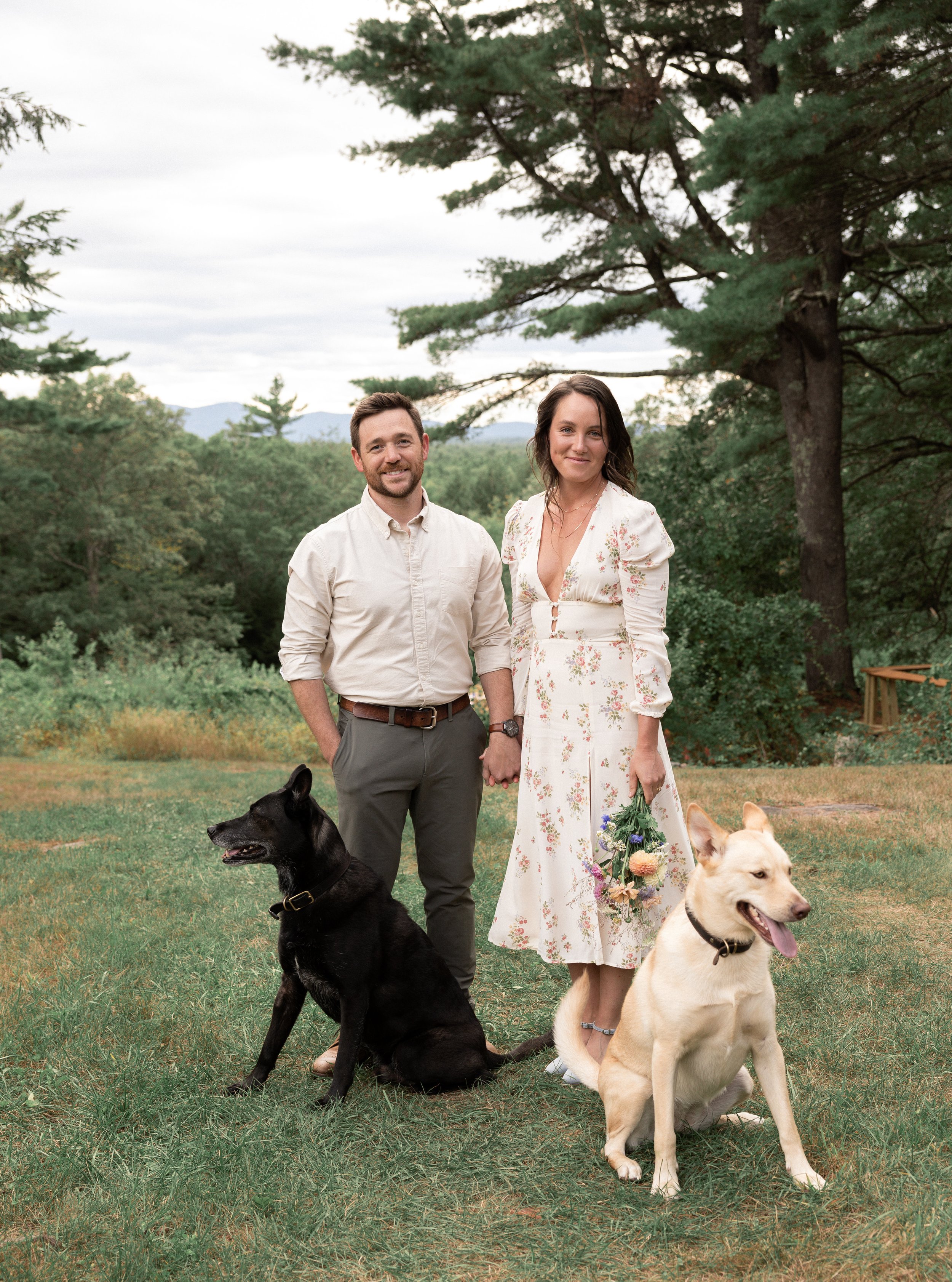 A couple holding hands, standing on grass outdoors, with two dogs. The woman is holding a bouquet of flowers, and is wearing a white dress with floral patterns. The man is dressed in pants and a light shirt. One dog is sitting next to the man, and the other is sitting next to the woman.