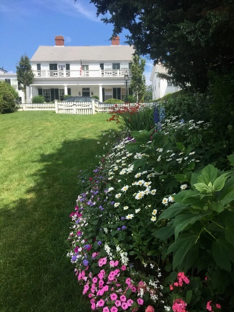 A white house with a front porch and red brick chimneys, set behind a well-maintained lawn and colorful flower bed, under a blue sky with a few clouds.