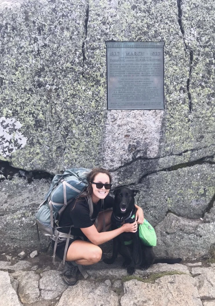 Woman wearing sunglasses crouching next to a black dog on a hike, both in front of a large rock with a bronze memorial plaque.