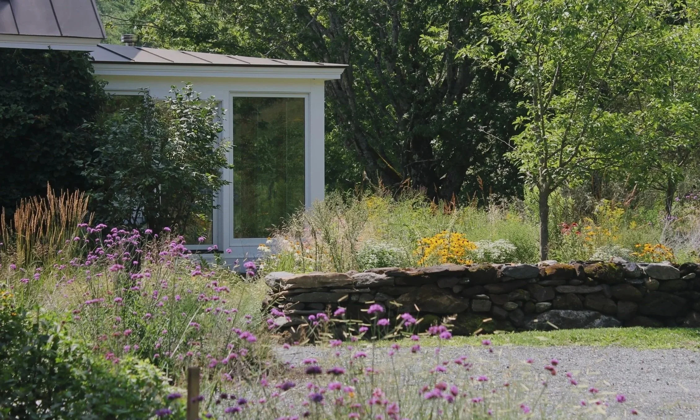 A garden with colorful flowers, a stone wall, green trees, and part of a house with a large window. Nautical-looking plants and a gravel pathway are visible.