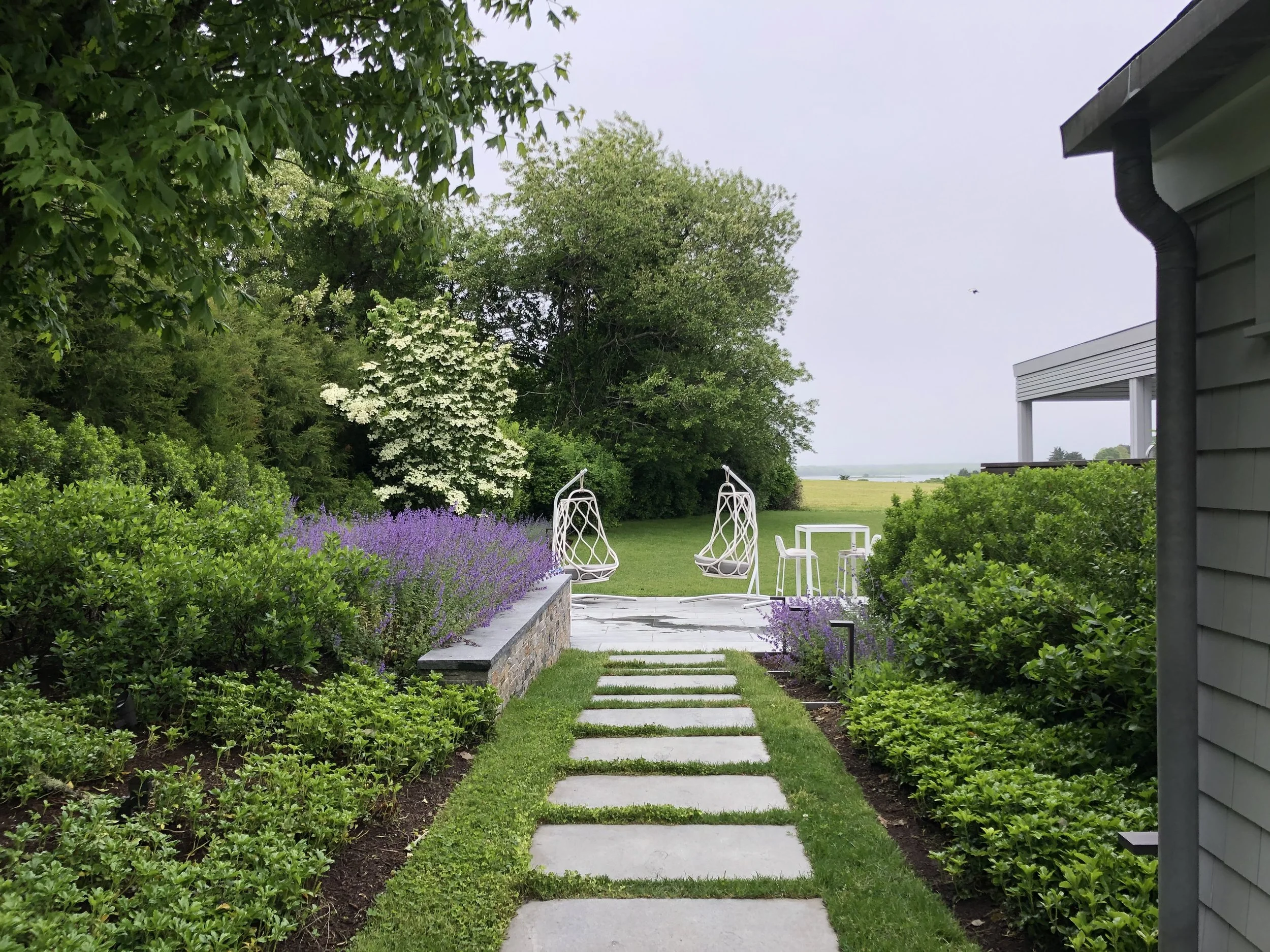 A garden path with stepping stones leading to a patio with two hanging chairs and a small table, surrounded by lush green bushes and flowers, with trees and an open field in the background.