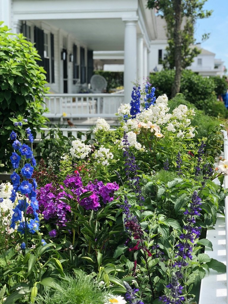 Colorful garden with purple, white, and blue flowers in front of a white house with a porch and column.