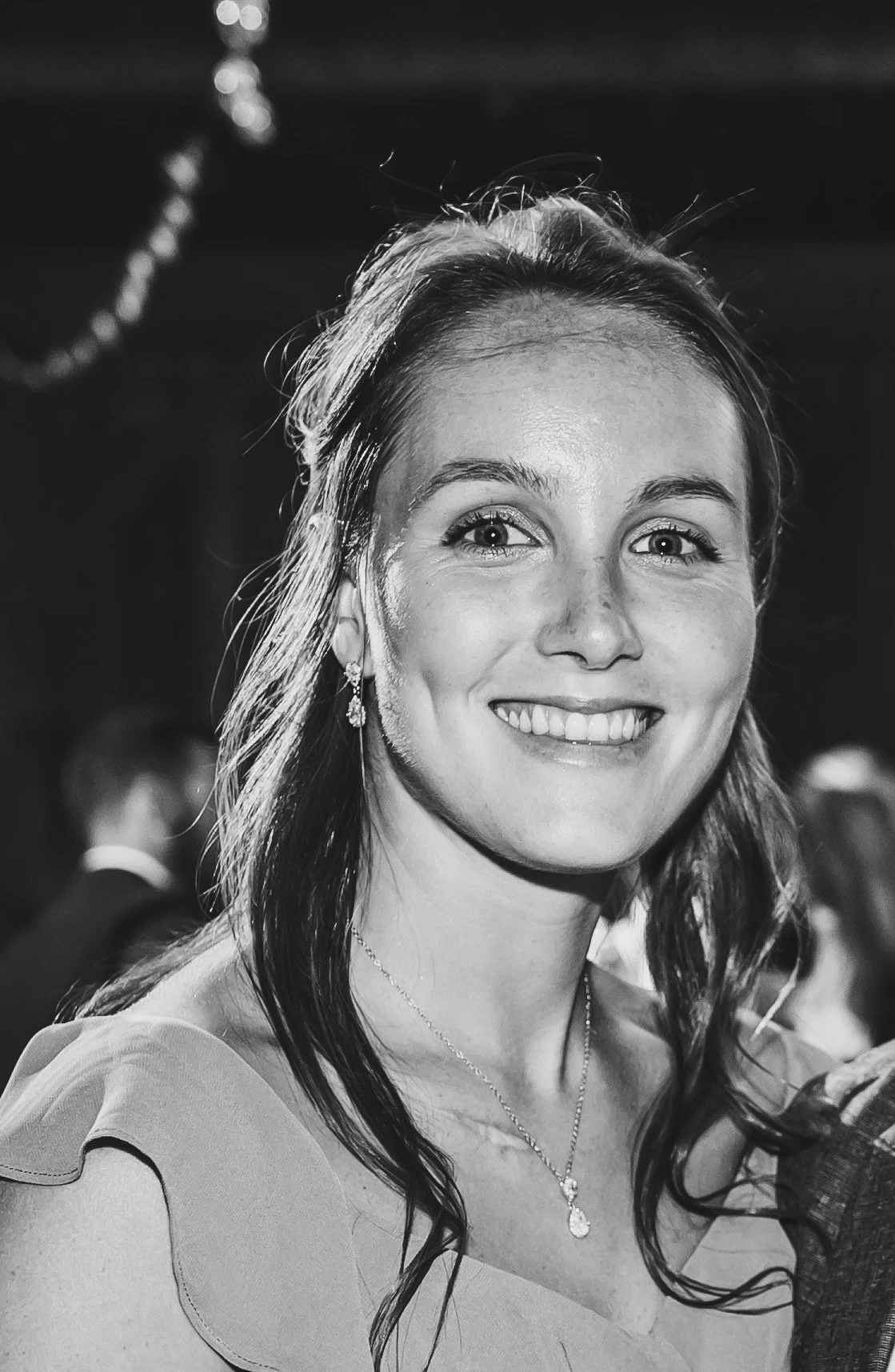 A smiling woman at a formal event, wearing earrings and a necklace, with people blurred in the background.