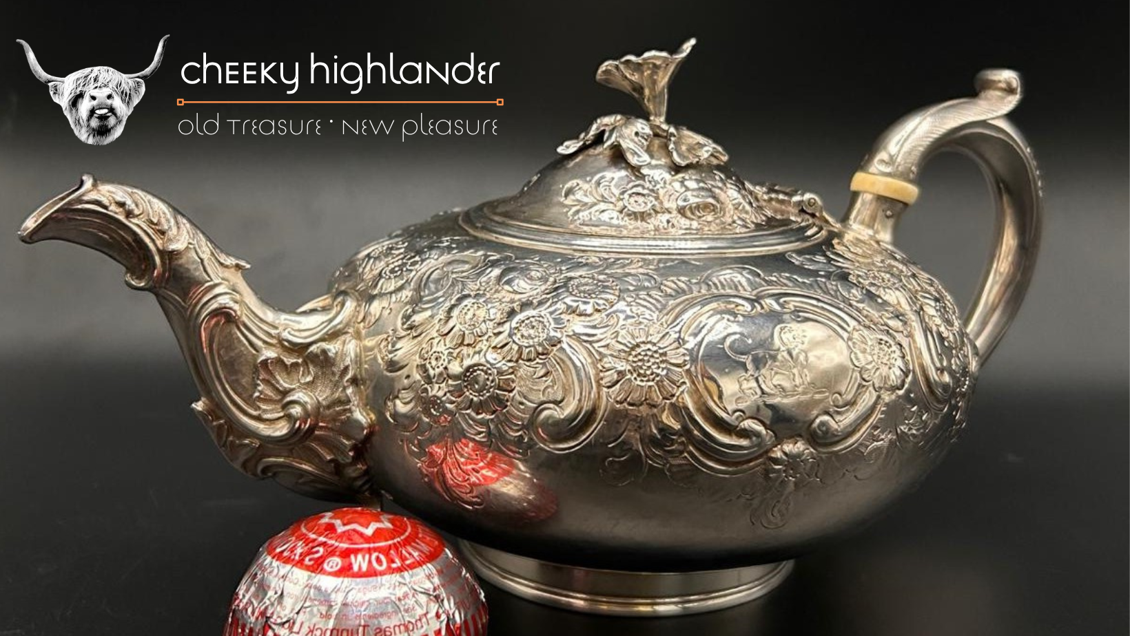 A silver ornate teapot with floral and animal engravings, placed on a black surface, with a Hershey's Kisses chocolate at the bottom left.