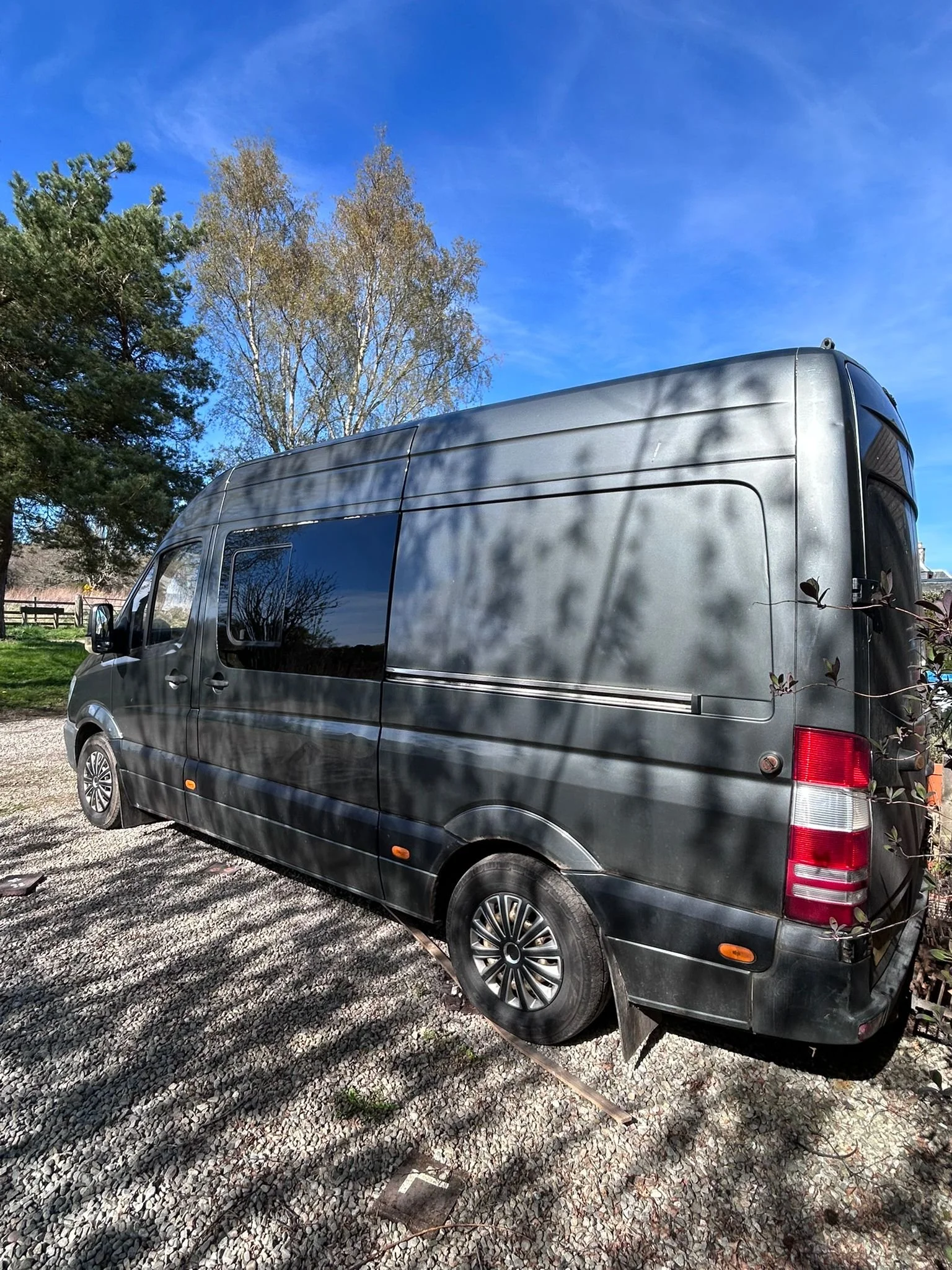 A black van parked on a gravel surface with trees and a blue sky in the background.