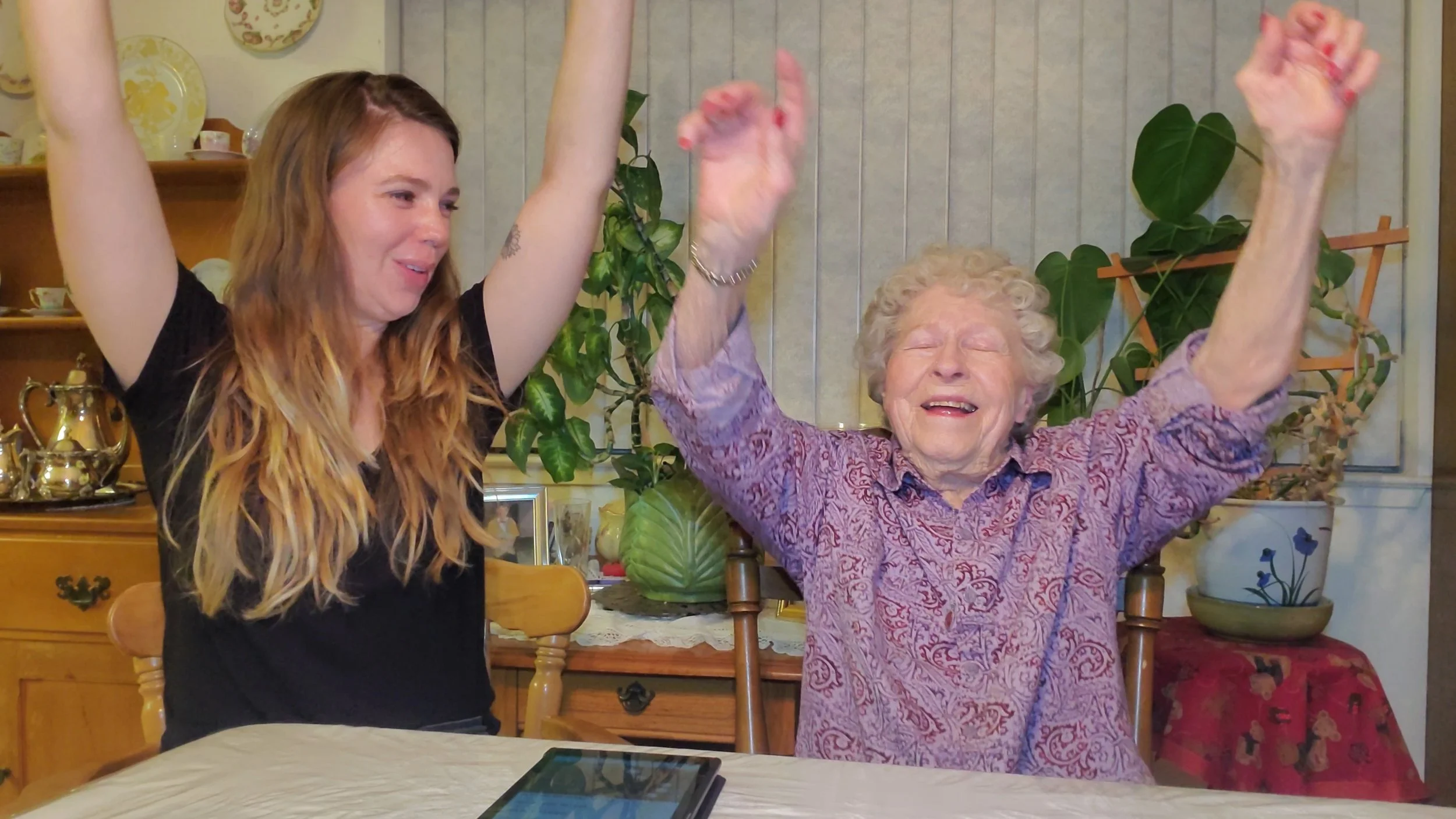 A young woman and an elderly woman sitting at a dining table, both with arms raised and smiling joyfully, in a cozy home with plants and decorative items in the background.