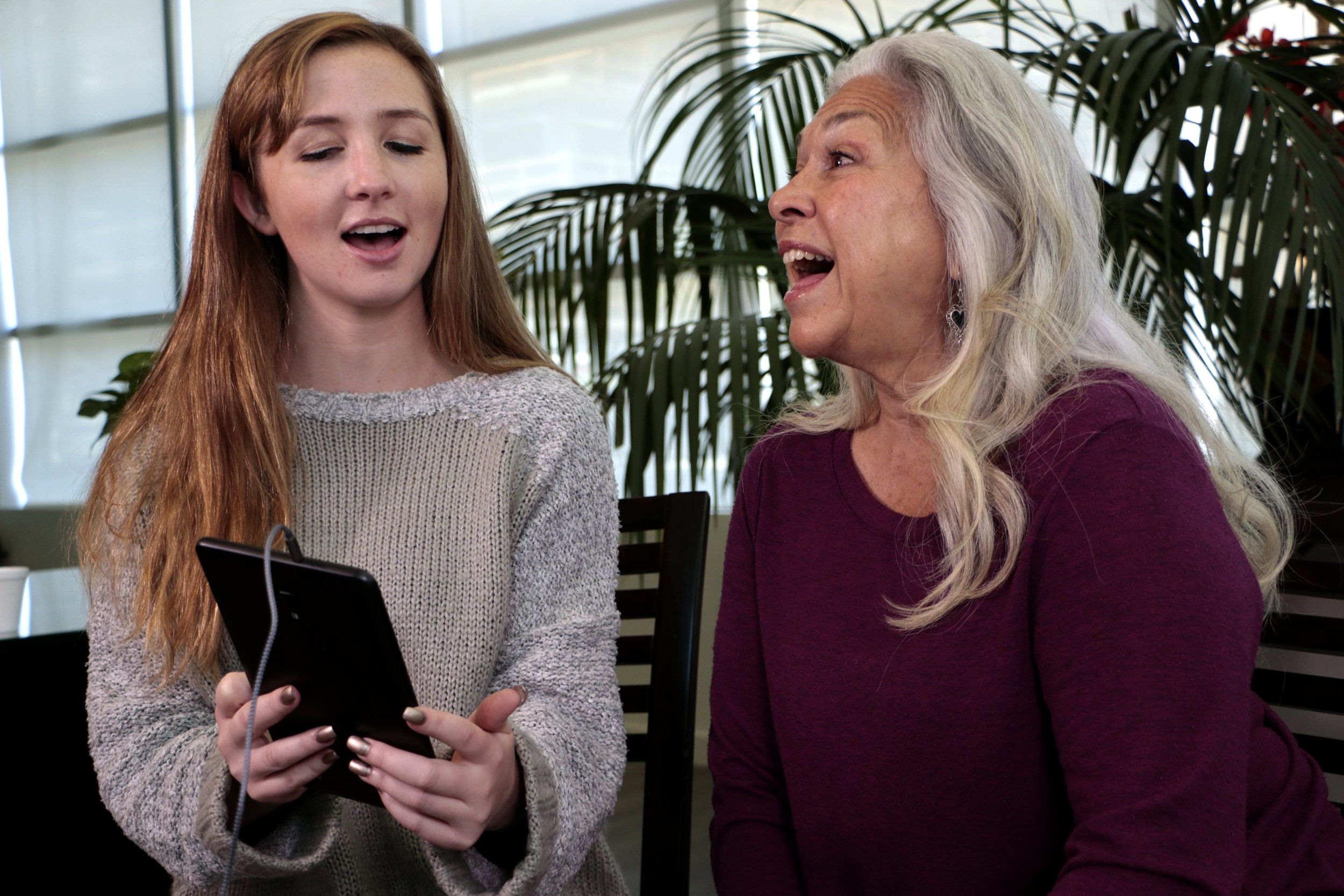 A young woman with long red hair and an elderly woman with long white hair are singing together while looking at a smartphone, indoors with plants in the background.