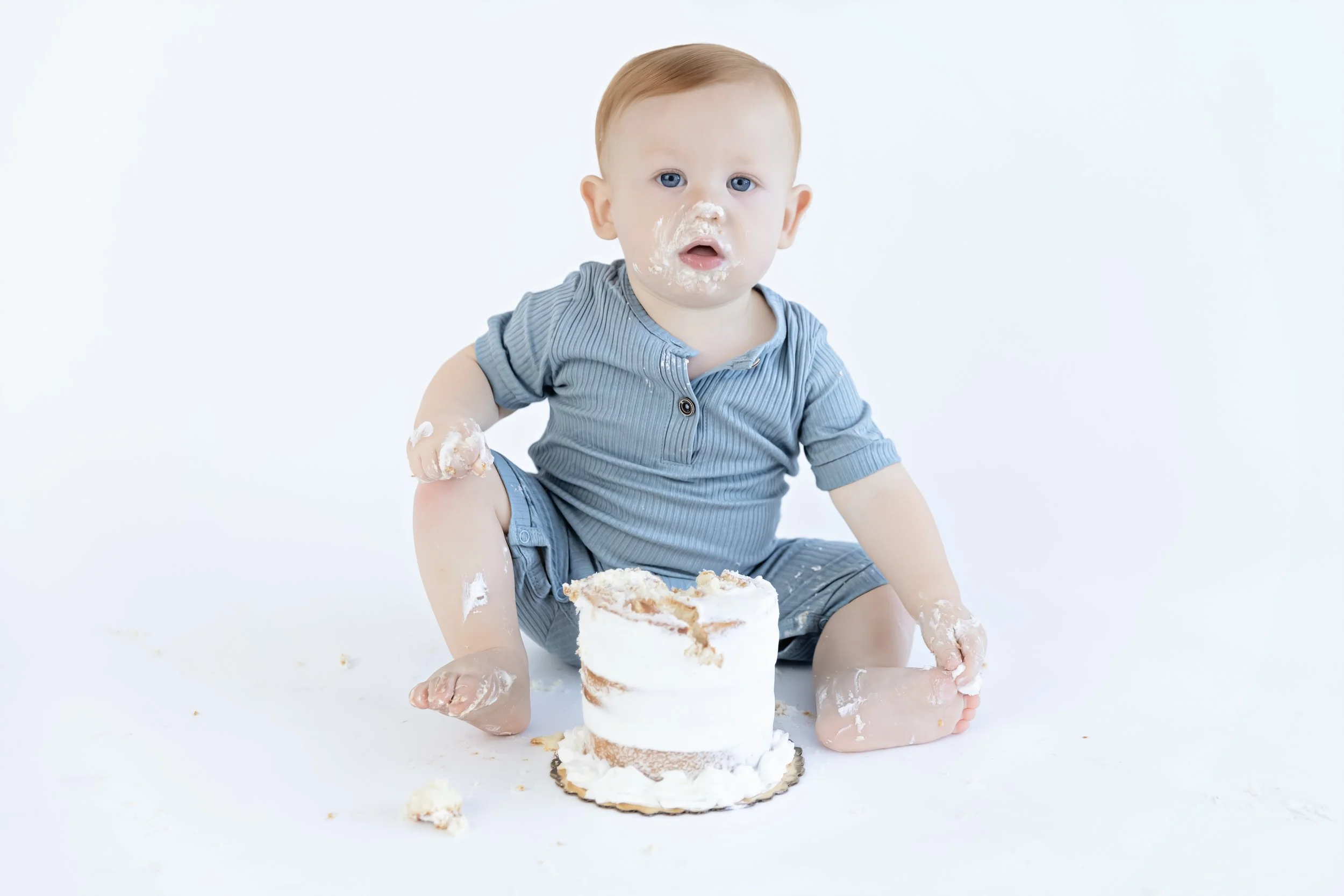 A smiling young girl with blue eyes and light brown hair sitting on a white floor with a small cake in front of her, in a bright, decorated room with white walls, plants, and rustic decor.