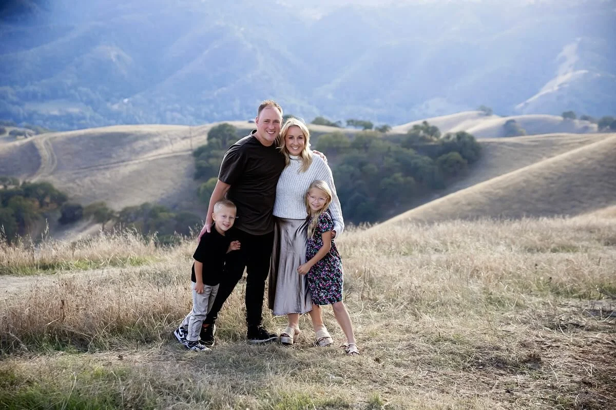 Happy family of four, two adults and two young children, posing outdoors on a sunny day amidst green trees and grass, smiling at the camera.