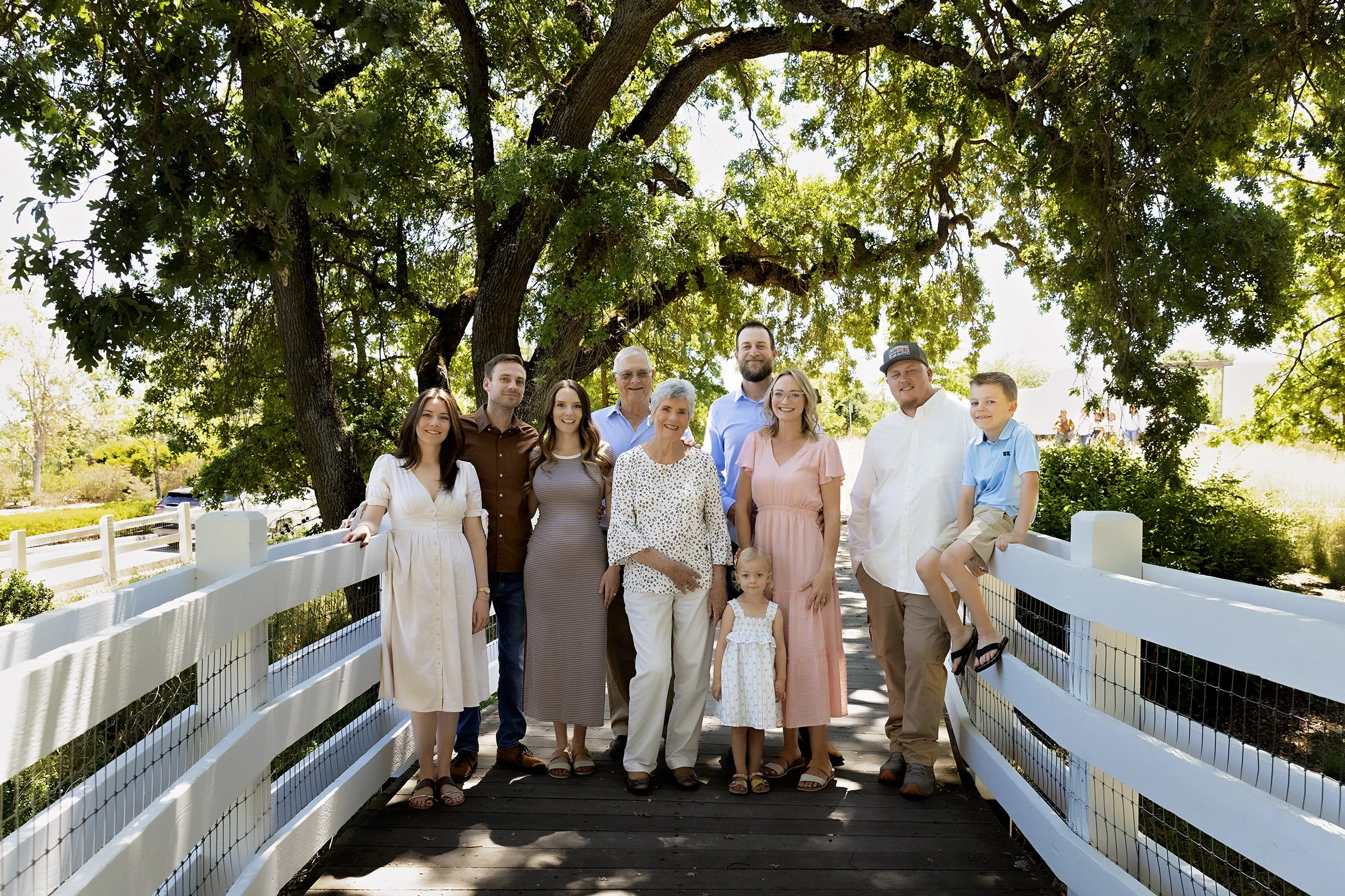A multi-generational family standing on a wooden bridge under a large tree, smiling for a group photo outdoors on a sunny day.