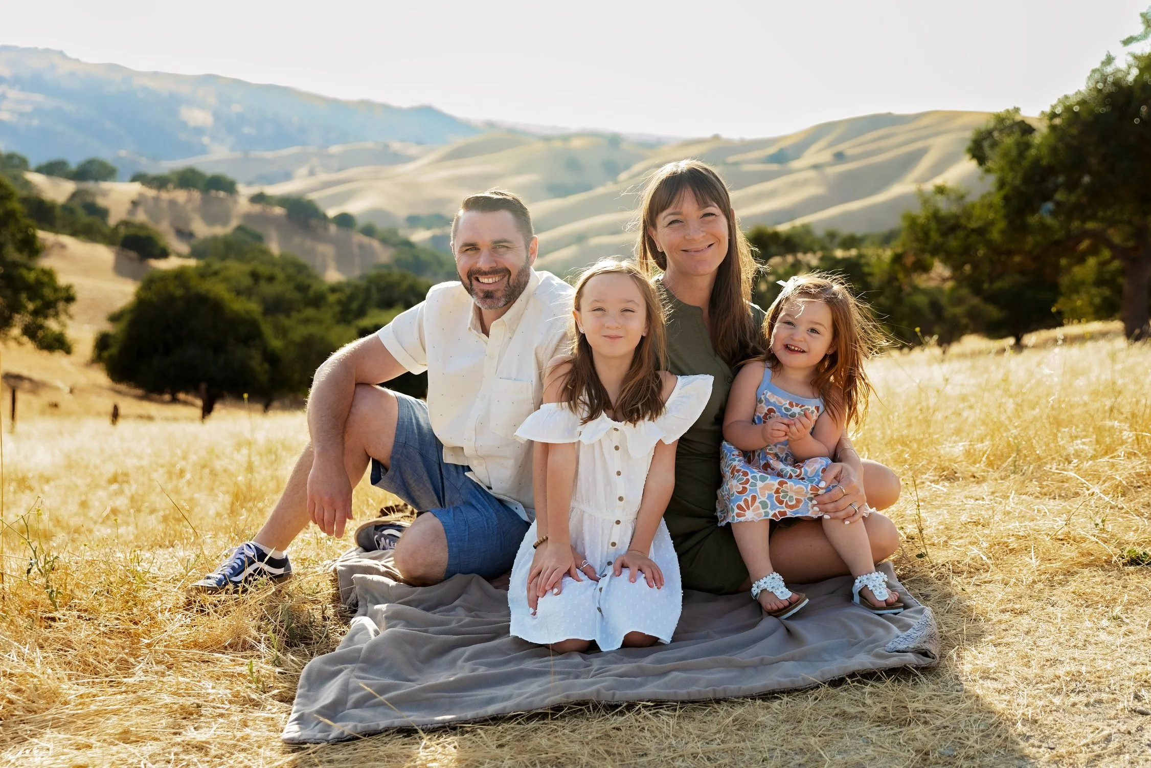 A happy family of four sitting on a blanket in a sunny, grassy field with rolling hills and trees in the background