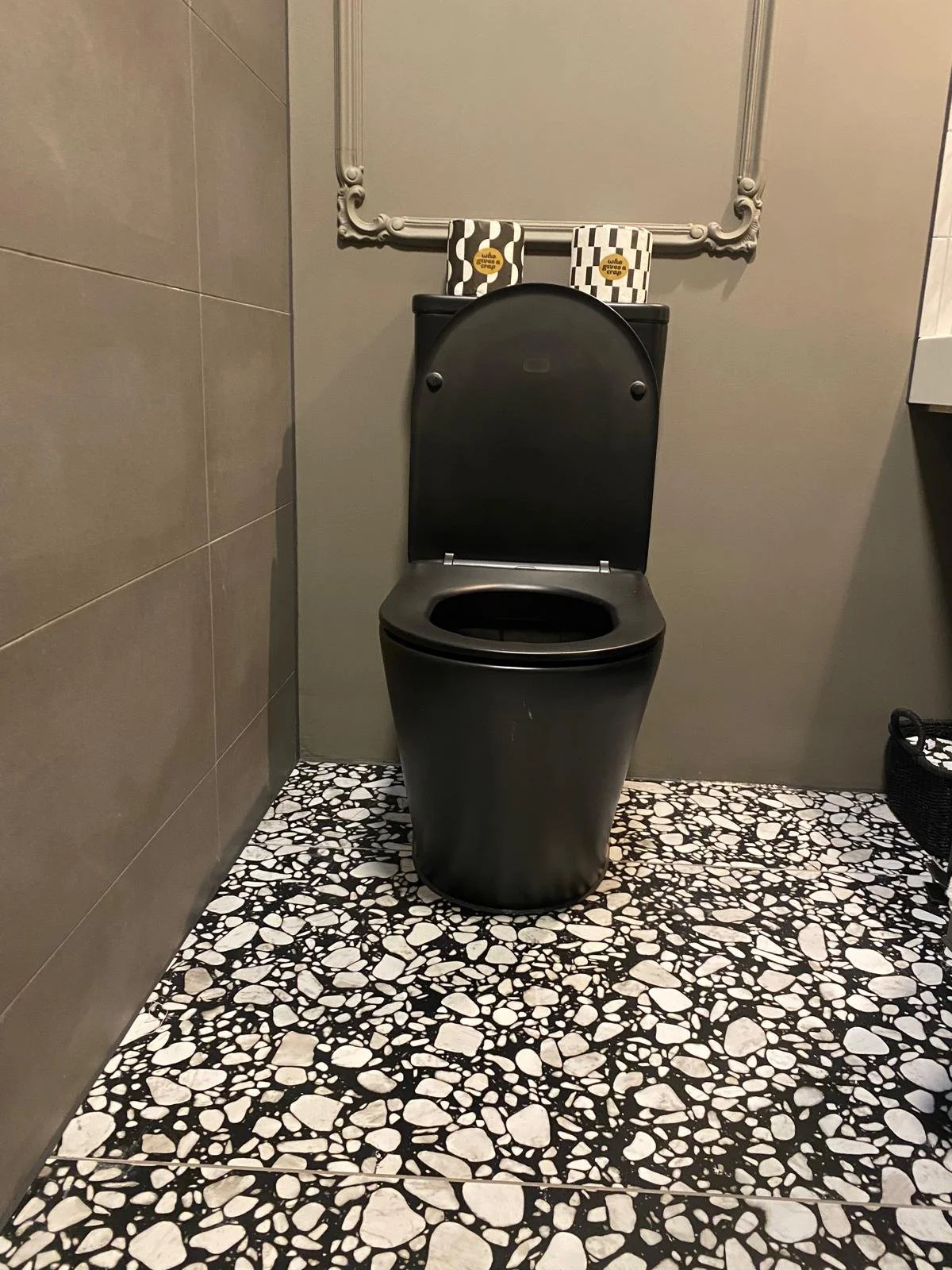 A black toilet with the lid up, placed on a black and white pebble-patterned floor in a bathroom. Behind it on the wall is a mirror and two boxes of cookies or snacks.