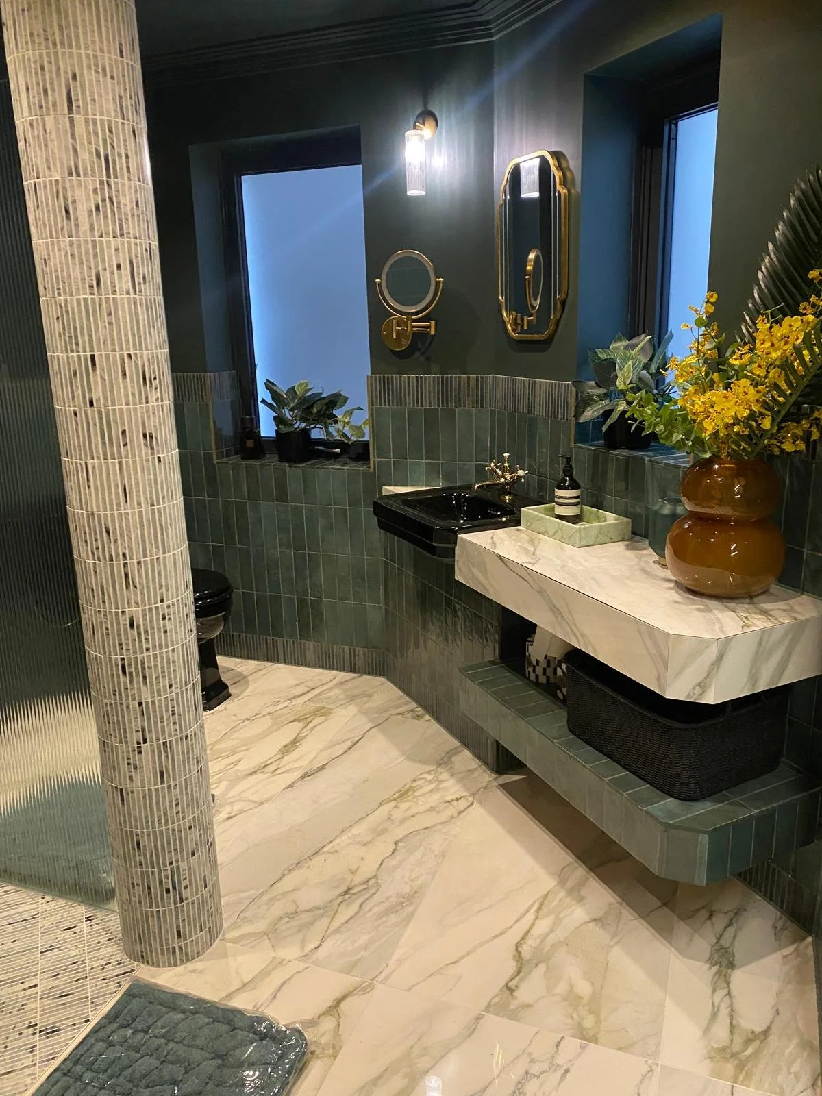 Modern bathroom with green tiled walls, marble floor, black sink, and decorative mirrors; includes potted plants and a vase with flowers.