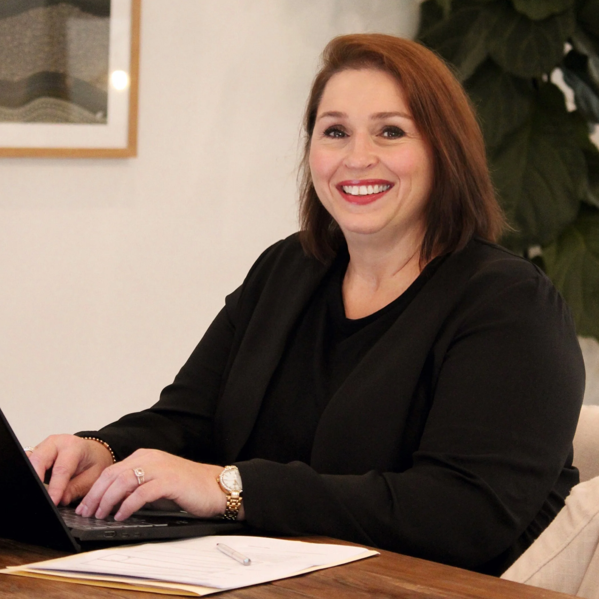 A woman with shoulder-length brown hair and wearing a black blazer is sitting at a table, smiling, with a laptop, notebook, and pen in front of her. Neilson and Co Legal - The Hunter Valley, Maitland and Newcastle.