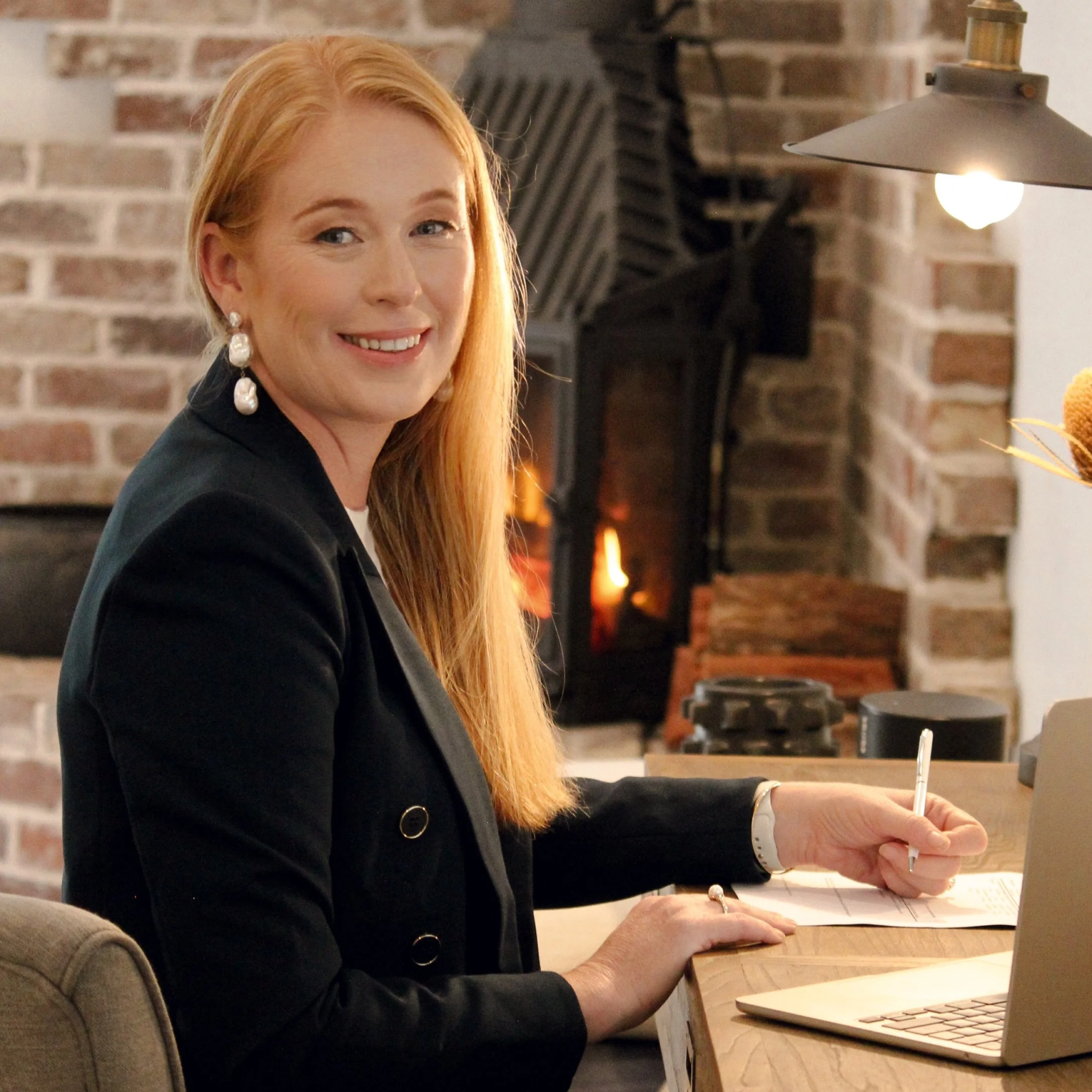 A woman with long red hair, wearing a black blazer and pearl earrings, sitting at a wooden table in front of a laptop, smiling, with a brick fireplace in the background. Louise Neilson - Neilson and Co Legal The Hunter Valley, Maitland and Newcastle